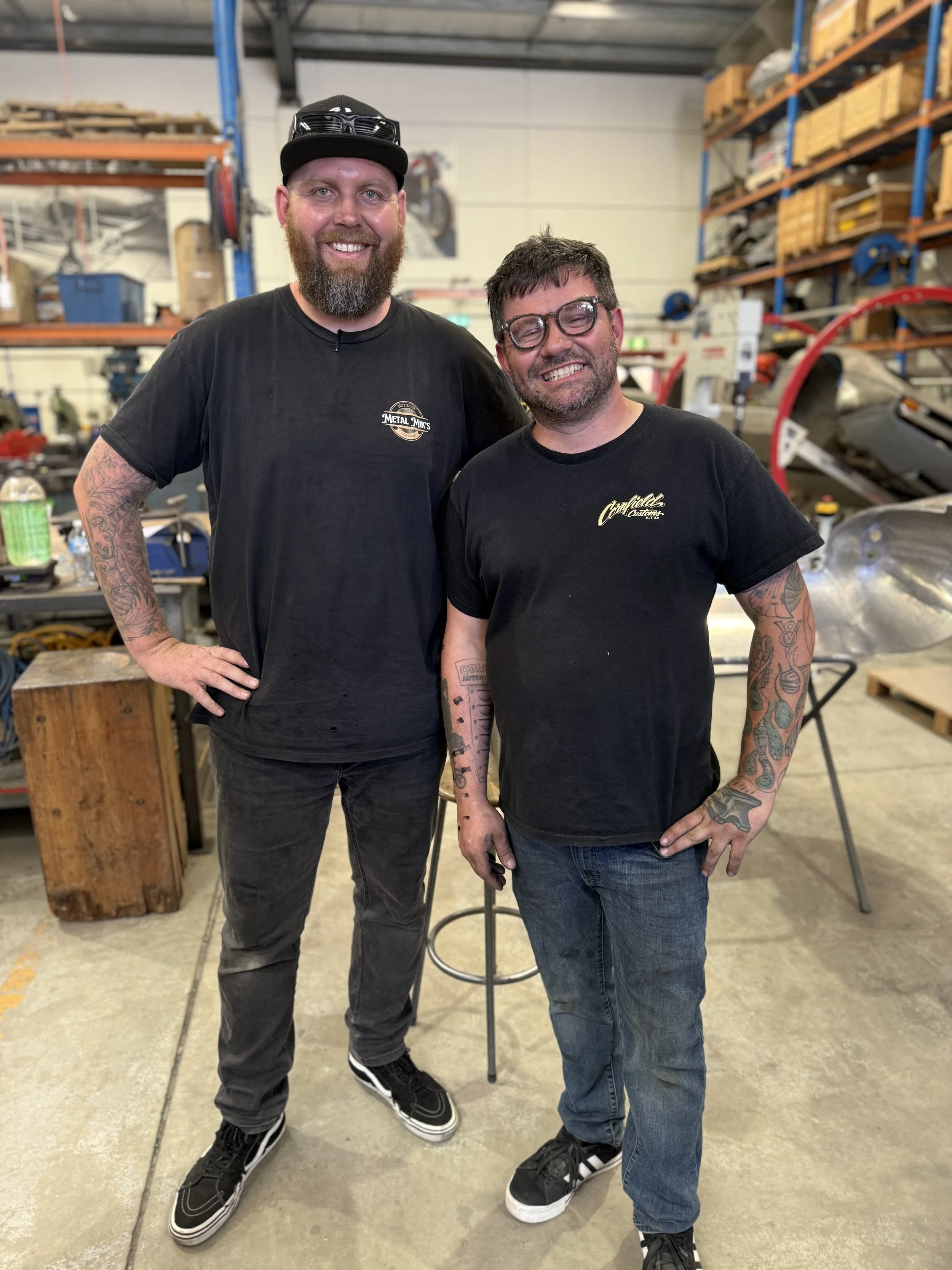 Two smiling men standing in a workshop, posing for a photo. The man on the left is tall with a beard, tattoos on his arms, and wearing a black cap and black t-shirt. The man on the right is shorter, wearing glasses, and a black t-shirt with tattoos on his arms. The background features metal shelves, workshop tools, and a partially assembled vehicle.