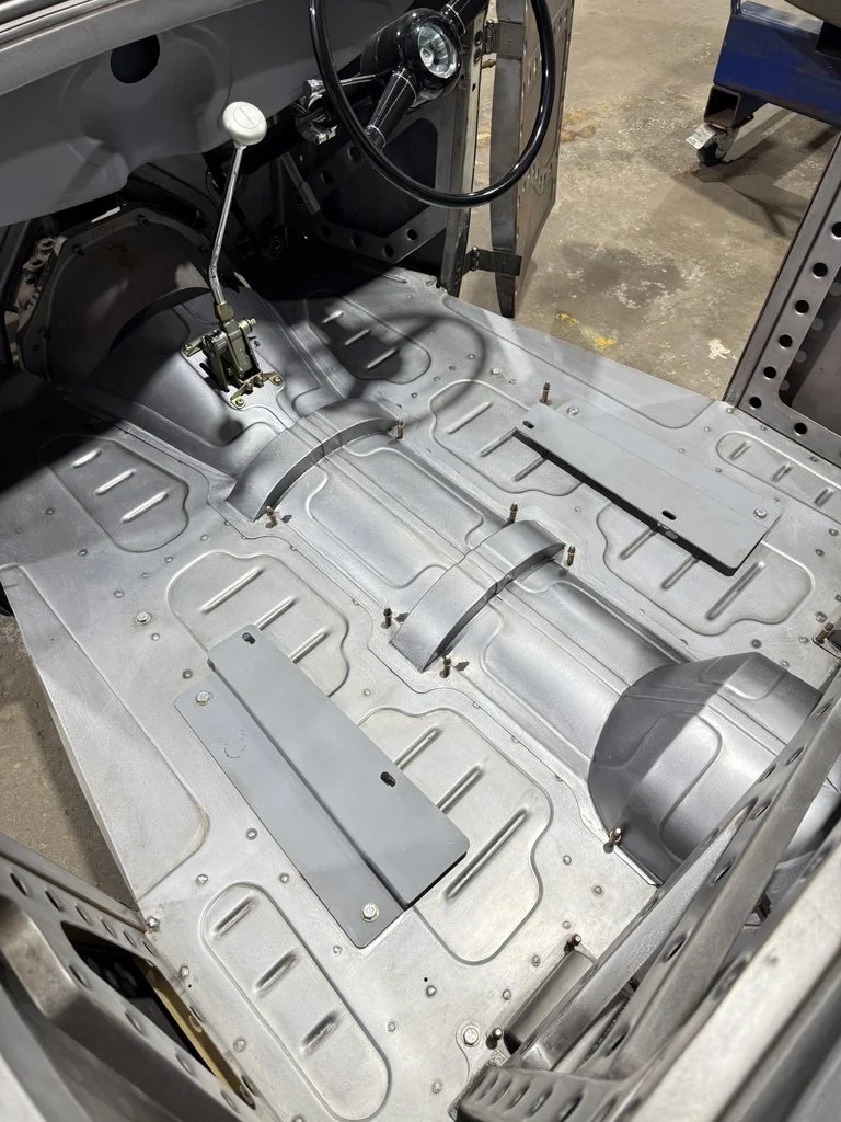 The interior of a vintage or classic car, showing the bare metal floor panels, some mounting brackets, and part of the steering wheel and dashboard.