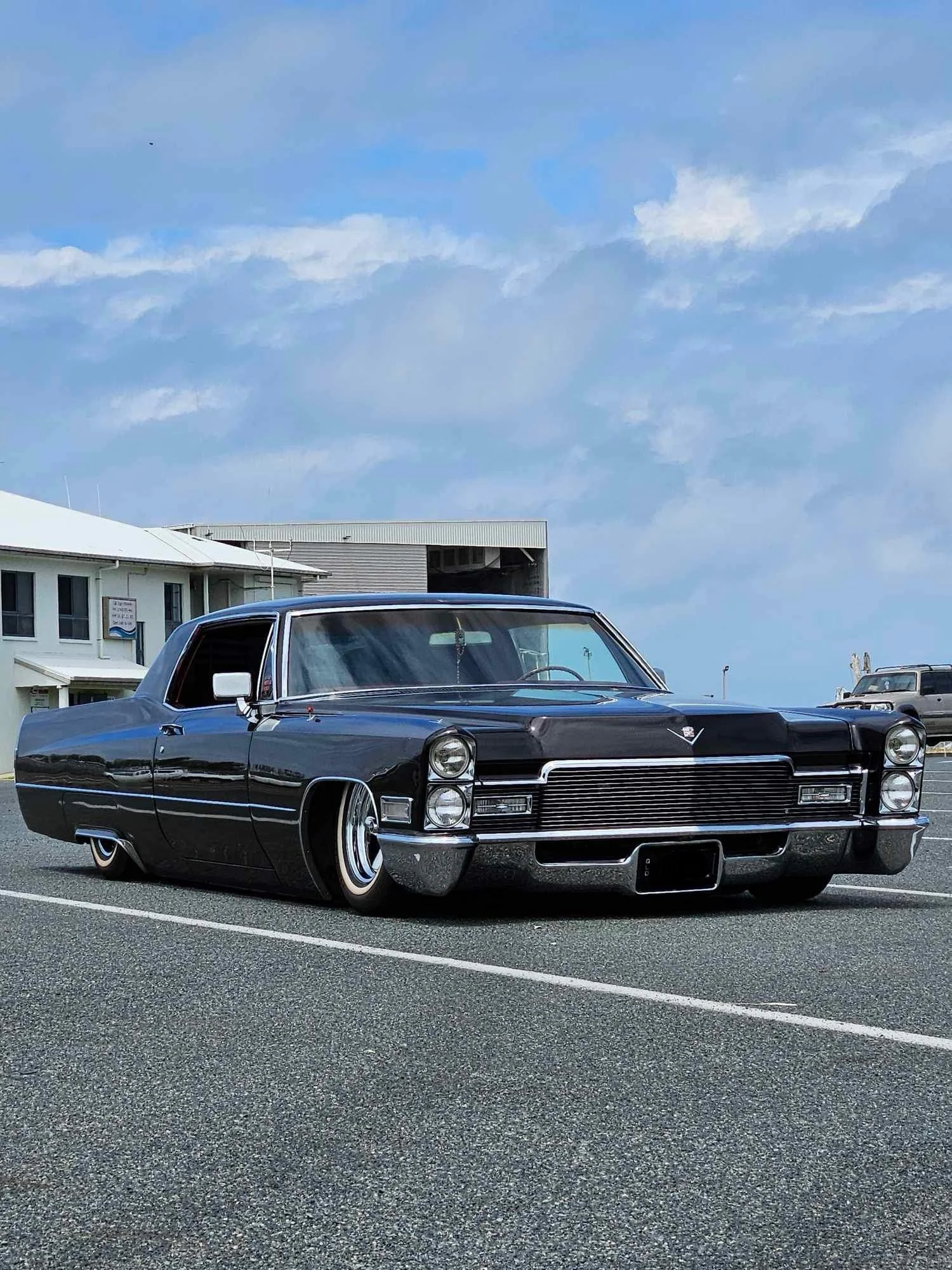 A vintage black Cadillac car parked in a parking lot under a partly cloudy sky.