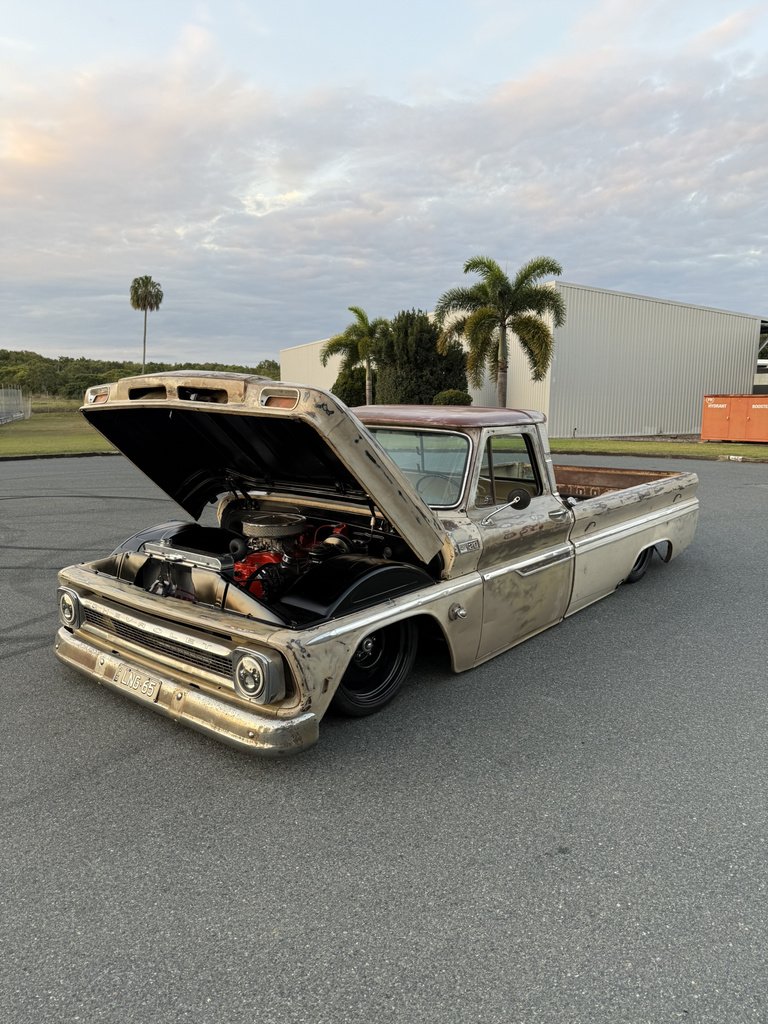 A vintage, rusty pickup truck with an open hood showing a red engine. The truck is parked on a paved surface with palm trees and industrial buildings in the background under a cloudy sky.