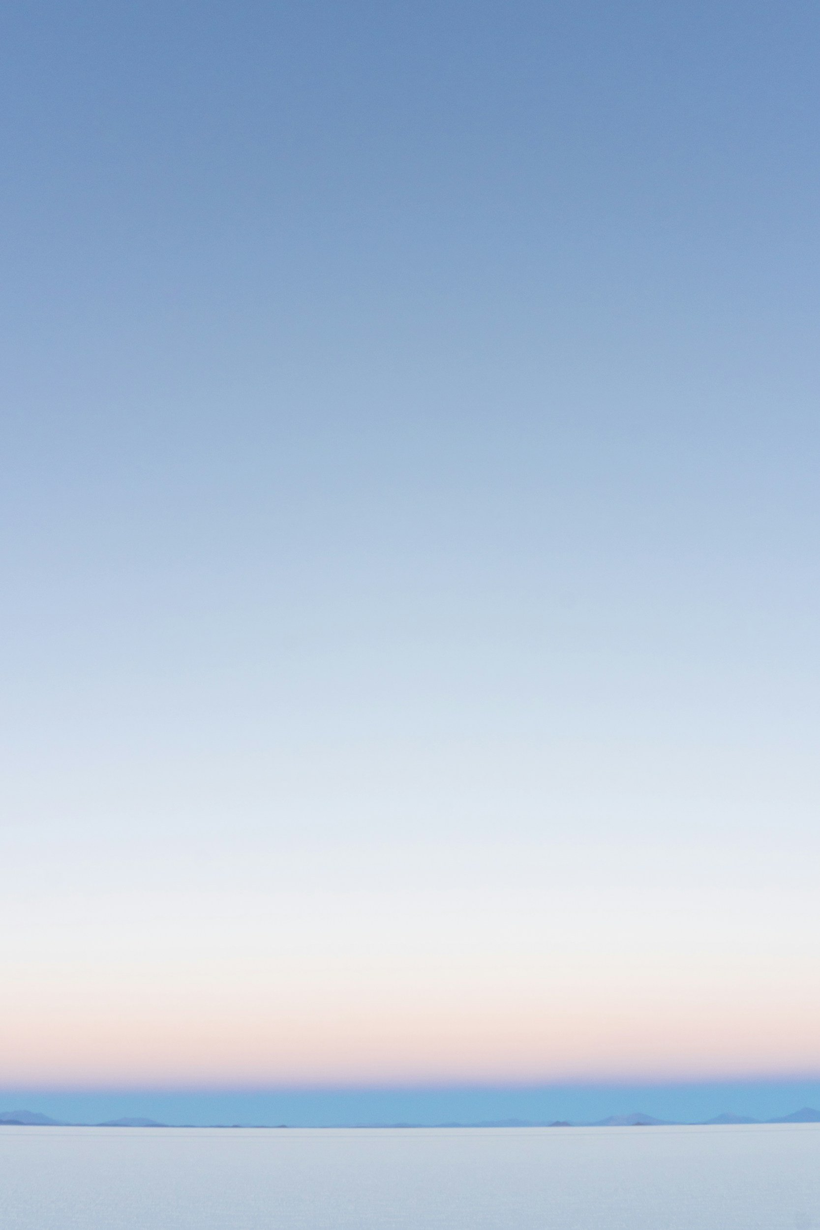 A serene landscape of a large body of water with mountains in the distance under a pastel-colored sky during dawn or dusk.