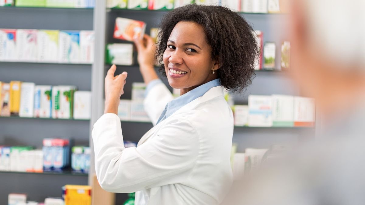 A smiling female pharmacist in a white lab coat stocks shelves of medicine in a pharmacy.