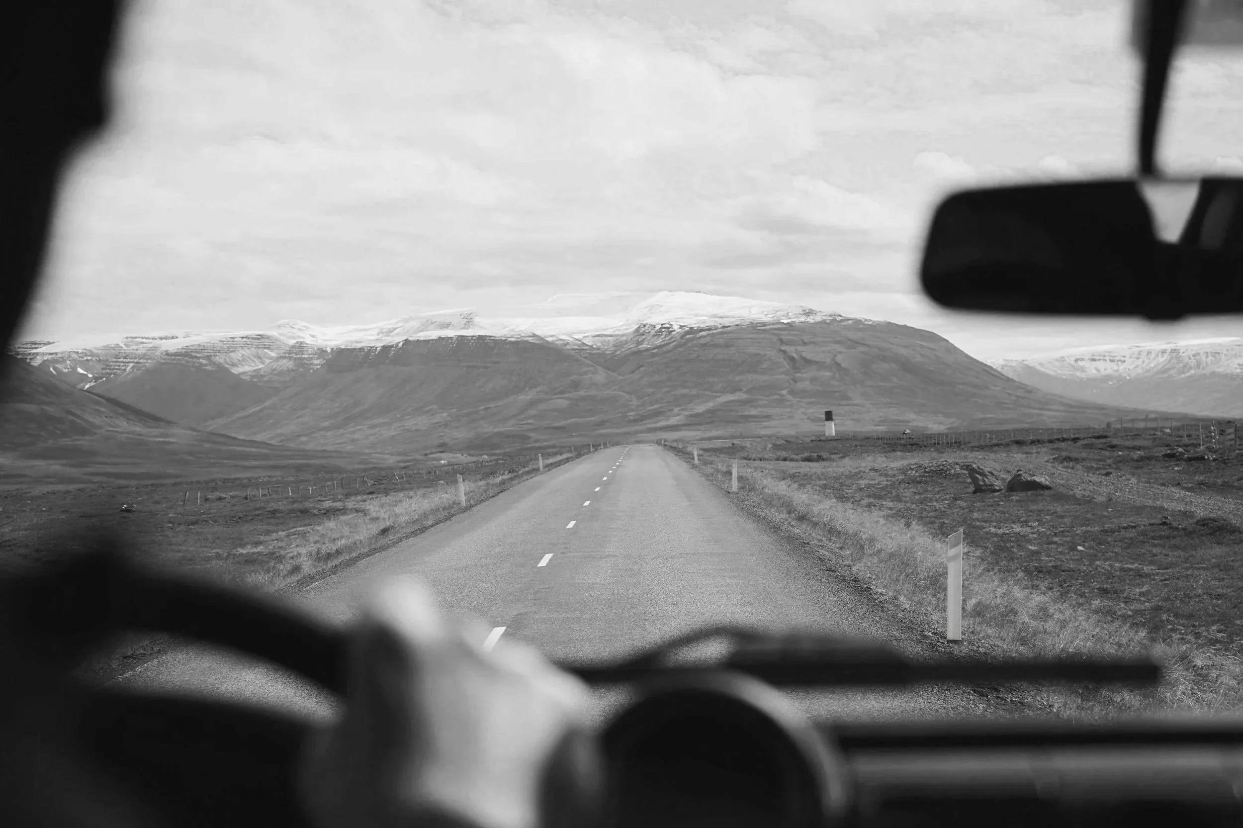 View of a long, empty rural road stretching towards distant mountains with snow on their peaks, seen from inside a vehicle with the dashboard and rearview mirror visible.
