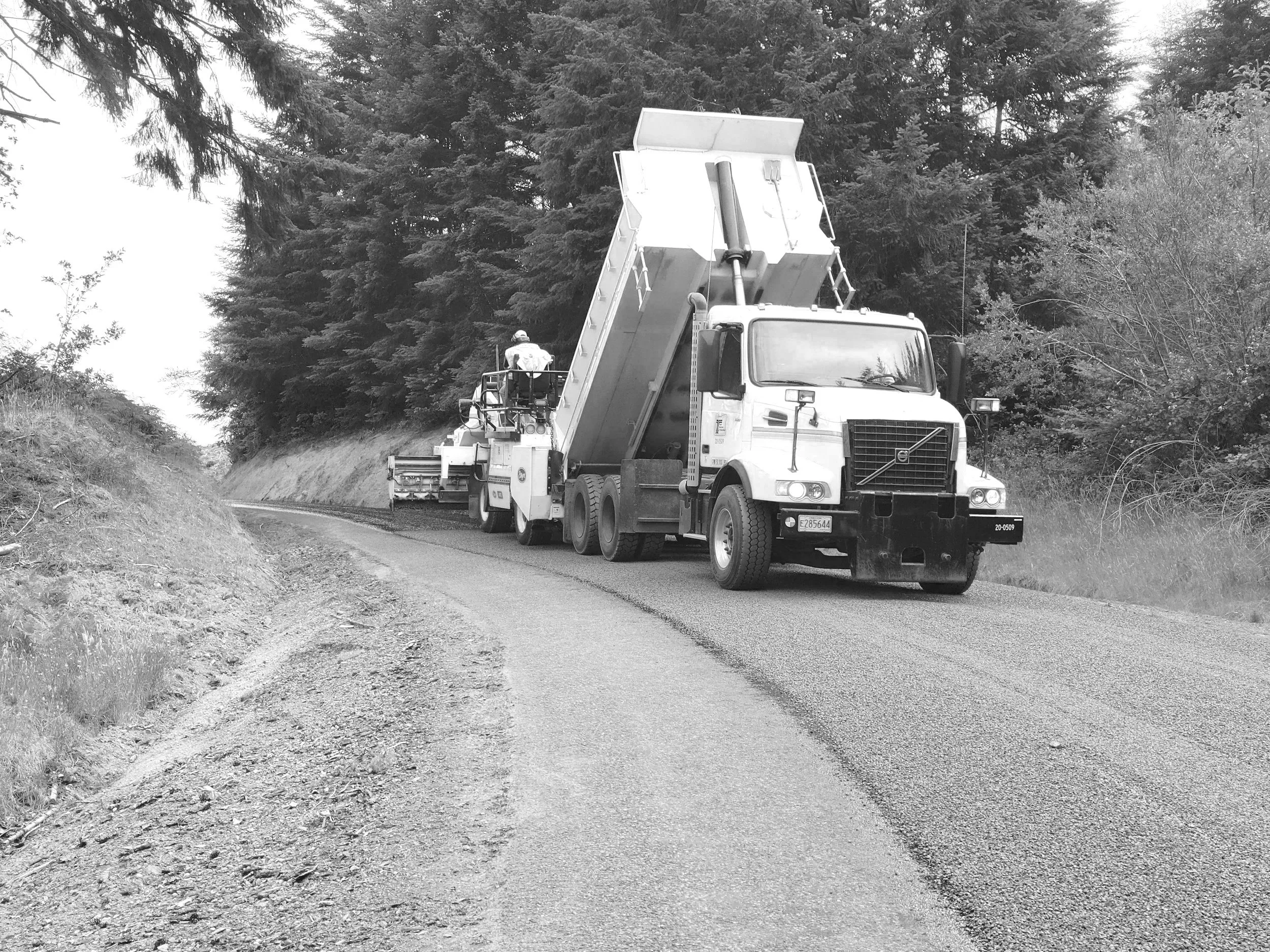 A road construction scene with a large dump truck and road paving equipment working on a curved, rural road surrounded by trees.