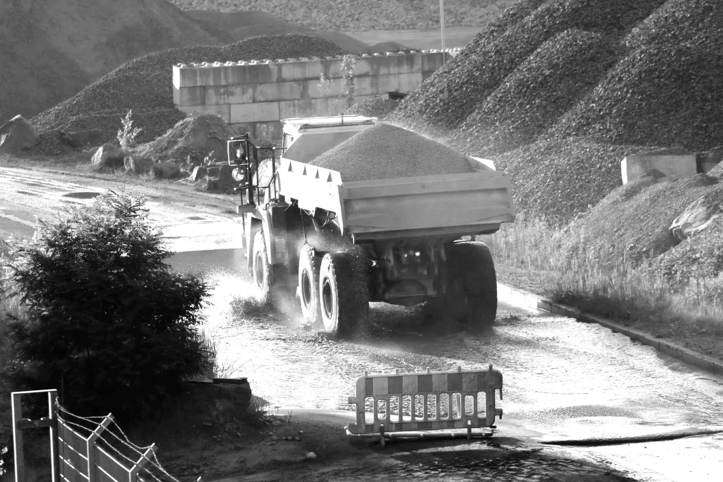 A large dump truck driving on a wet road carrying a load of gravel, with mountains of gravel and dirt on either side of the road.
