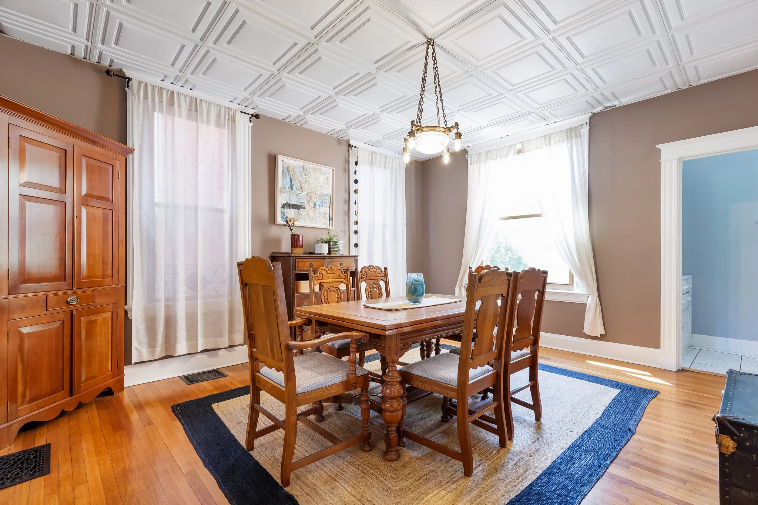 A wooden dining table with six matching chairs in a well-lit room with hardwood floors, cream-colored walls, large windows with white curtains, a wooden cabinet, a framed painting, and a chandelier hanging from a decorative ceiling.
