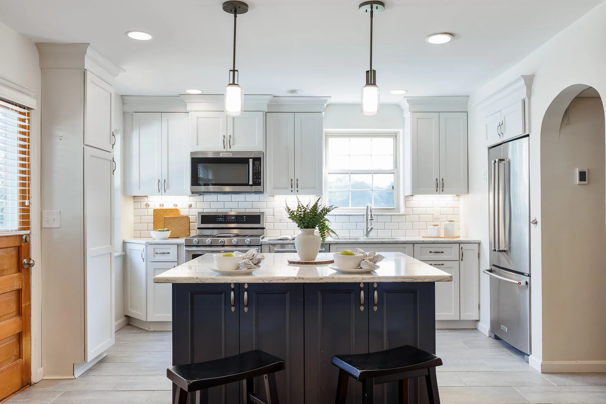 A modern kitchen with white cabinets, a marble island, stainless steel appliances, pendant lights, a window above the sink, and a few bowls and plants on the counter.