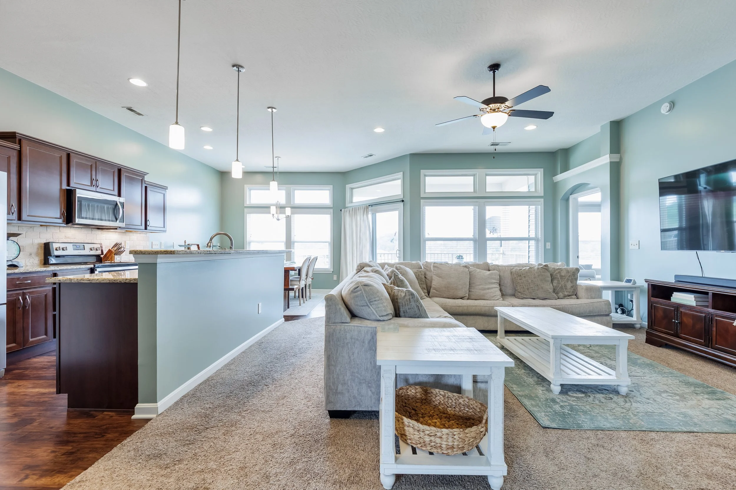 Open-concept living room and kitchen with light green walls, beige carpet, large windows, ceiling fan, and modern furniture.