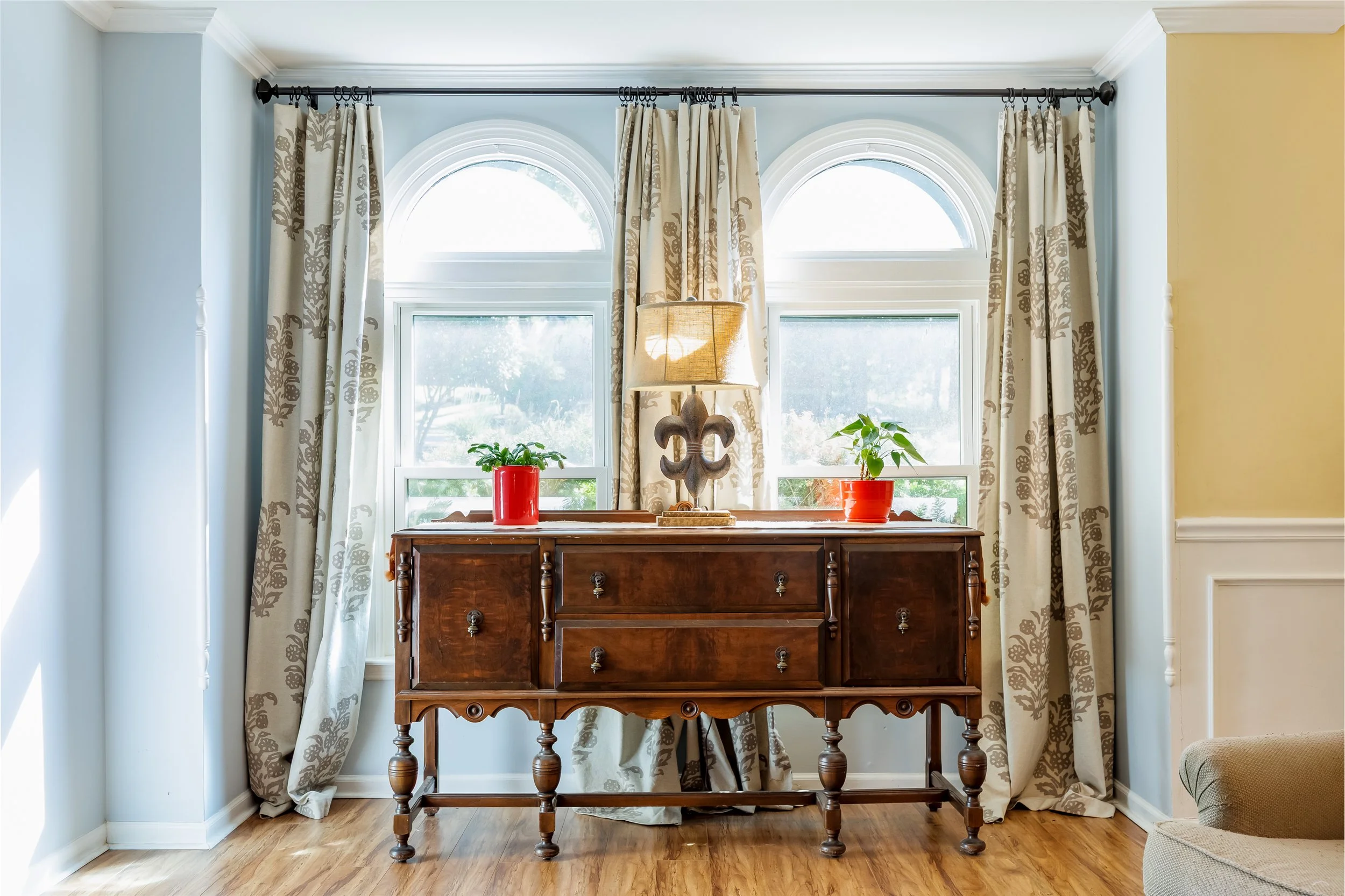Interior of a room with a wooden sideboard, two potted plants in red pots on top, a decorative lamp in the middle, and patterned curtains hanging on a black curtain rod in front of two large arched windows, with light-colored walls and hardwood floor