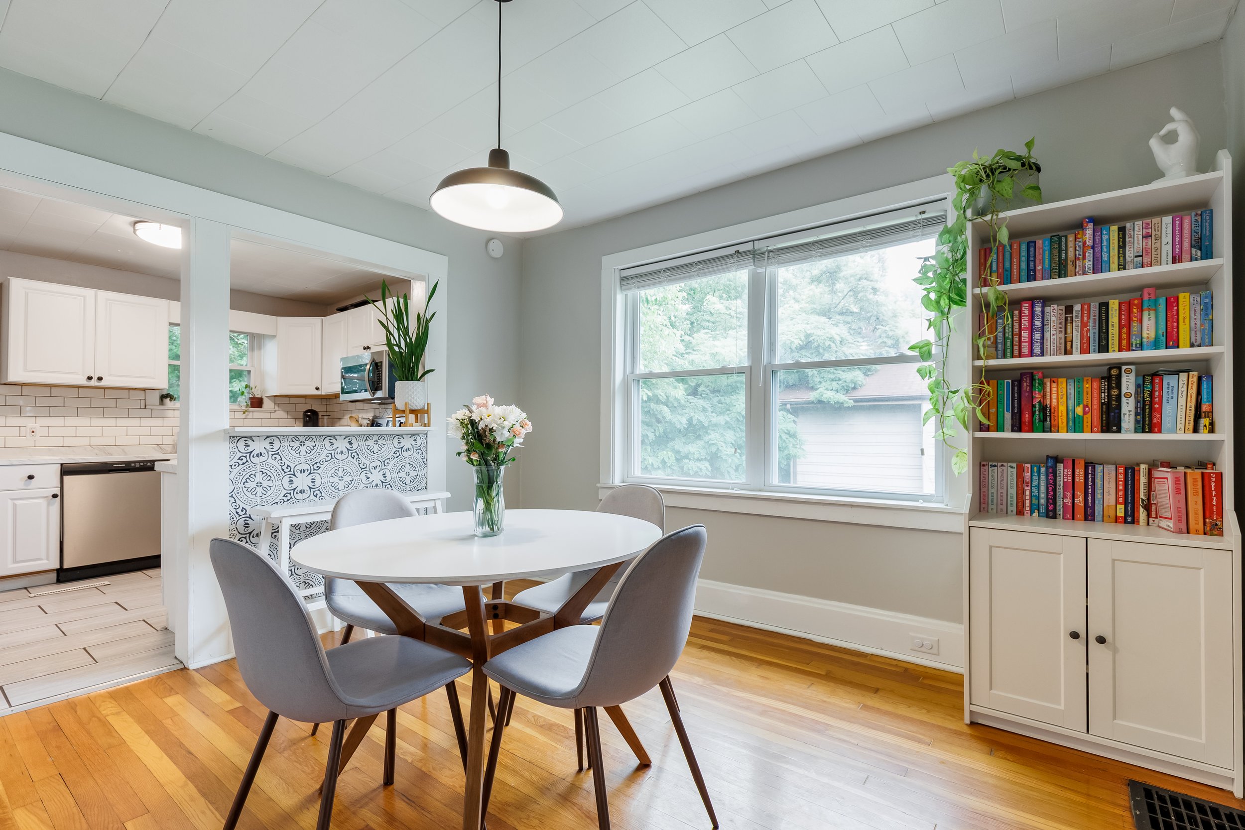 A bright dining area with a round white table surrounded by five gray chairs. A vase with flowers sits on the table. Large windows let in natural light, and a bookshelf filled with colorful books is next to the window. The kitchen with white cabinets