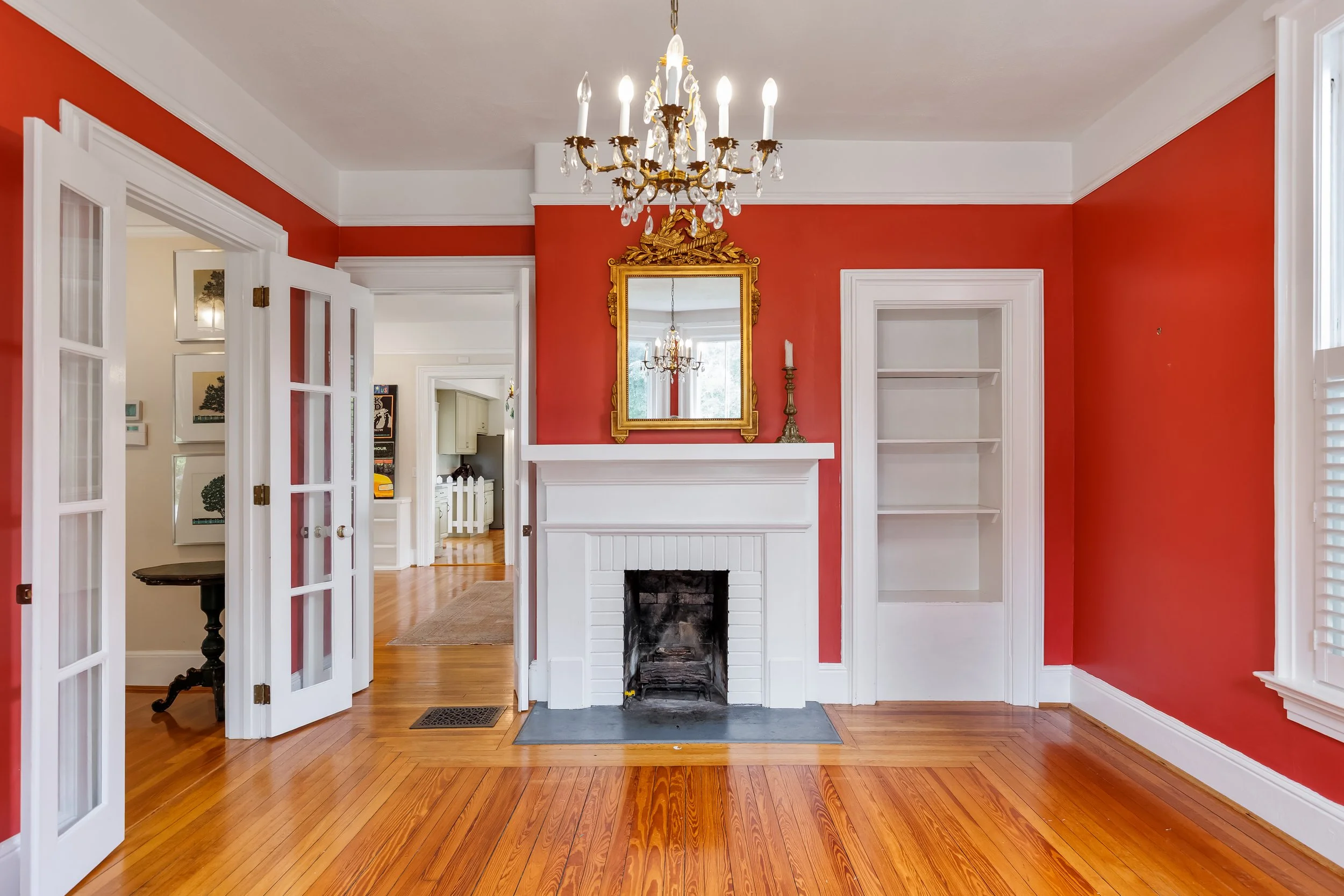 Interior of a room with red walls, white trim, a white fireplace with a gold ornate mirror above it, a chandelier hanging from the ceiling, and hardwood floors.