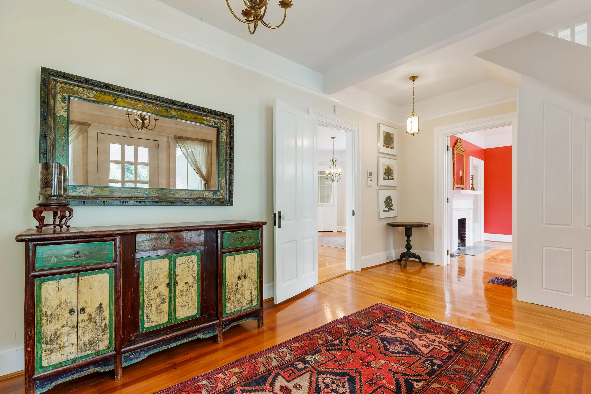 Interior view of a home with hardwood floors, a red and black patterned rug, a green and multicolored antique cabinet, wall art, and multiple ceiling light fixtures. A large mirror reflects a window with curtains.