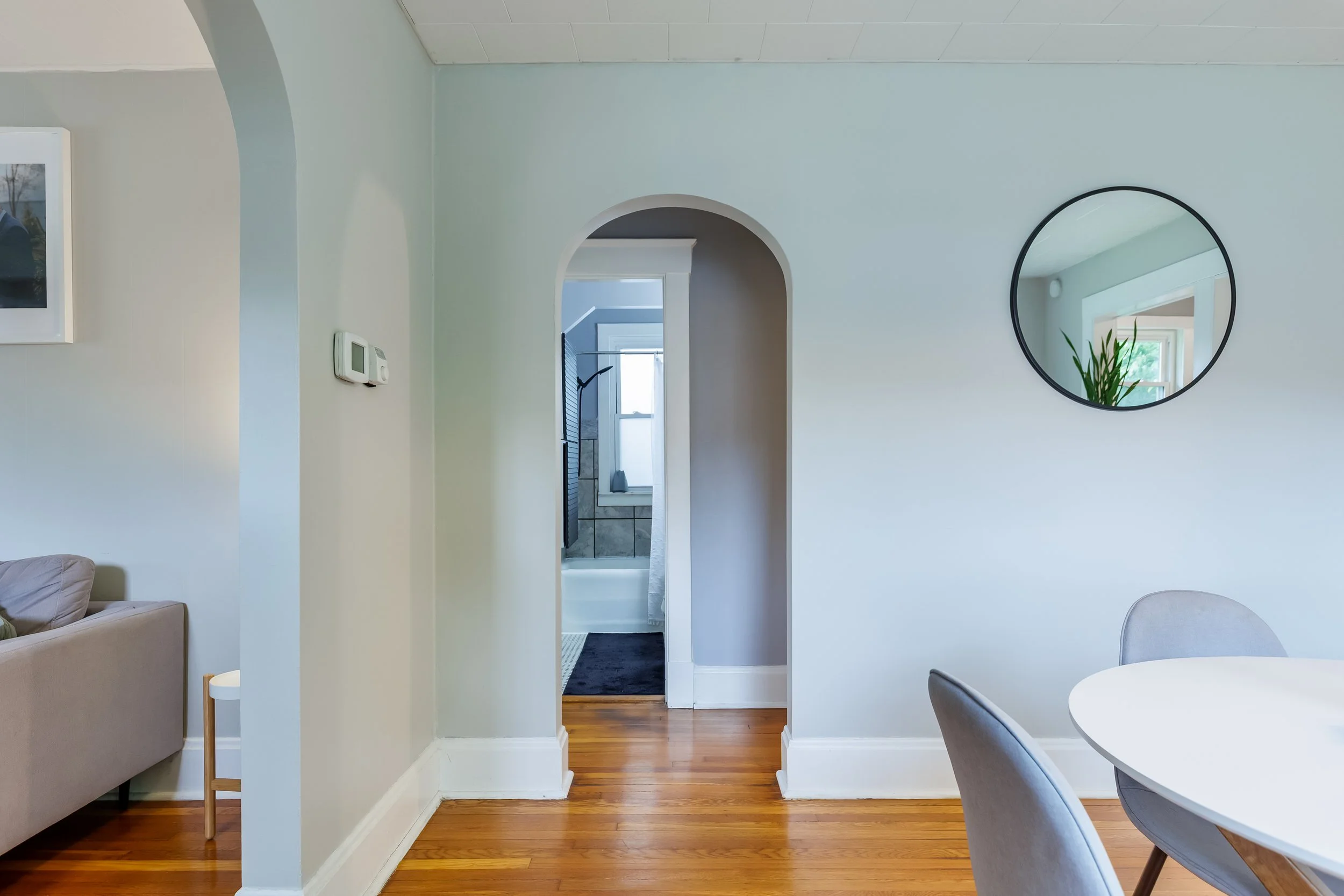 Living room with white walls, wood floor, and a doorway to a bathroom, which has a window and tiled walls. A round mirror hangs on the wall and reflects a window with greenery outside. There is a beige couch on the left and a white round table on the