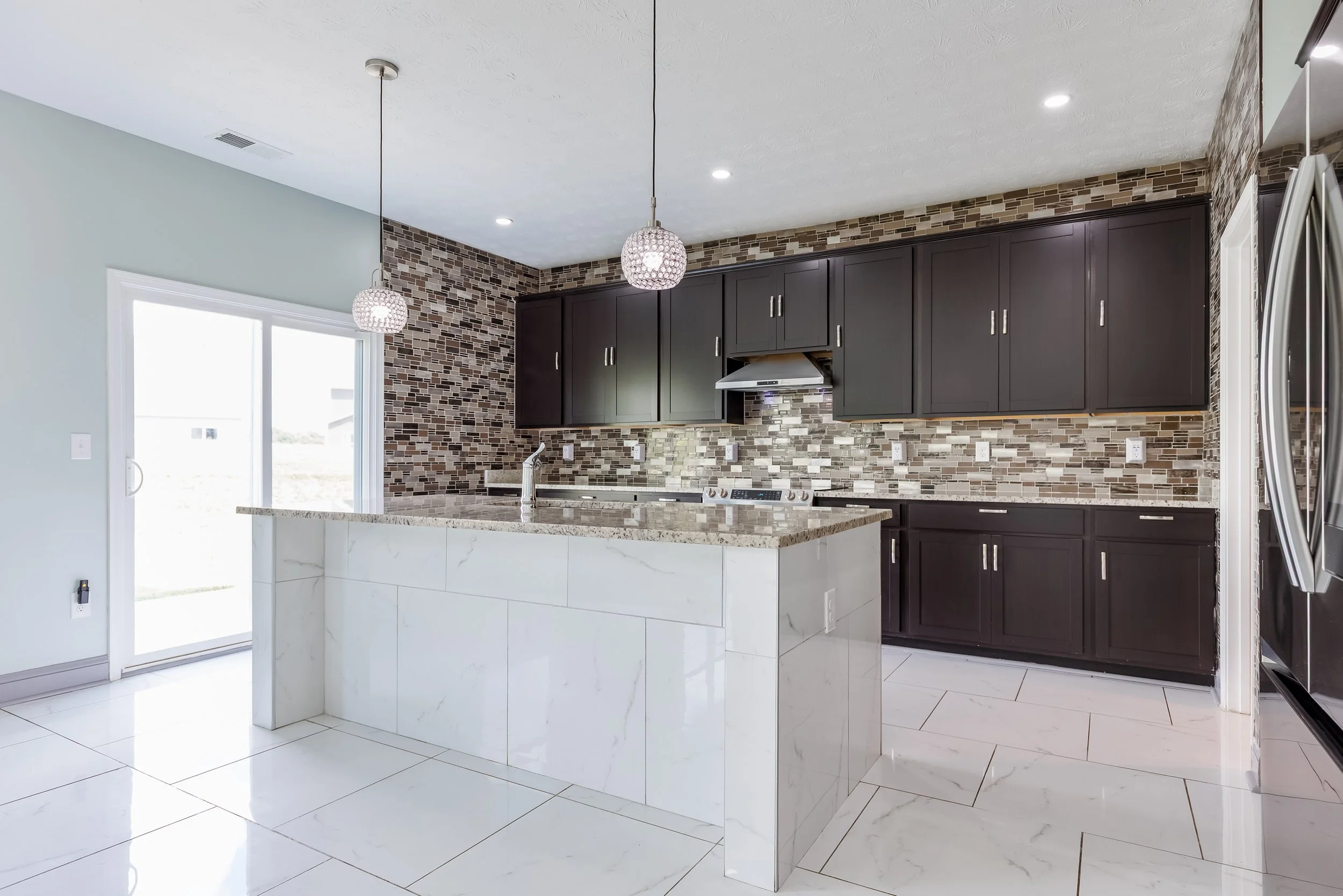 Modern kitchen with white tiled floor, black cabinets, mosaic tile backsplash, white kitchen island, stainless steel range hood, two hanging pendant lights, sliding glass door, and stainless steel refrigerator.