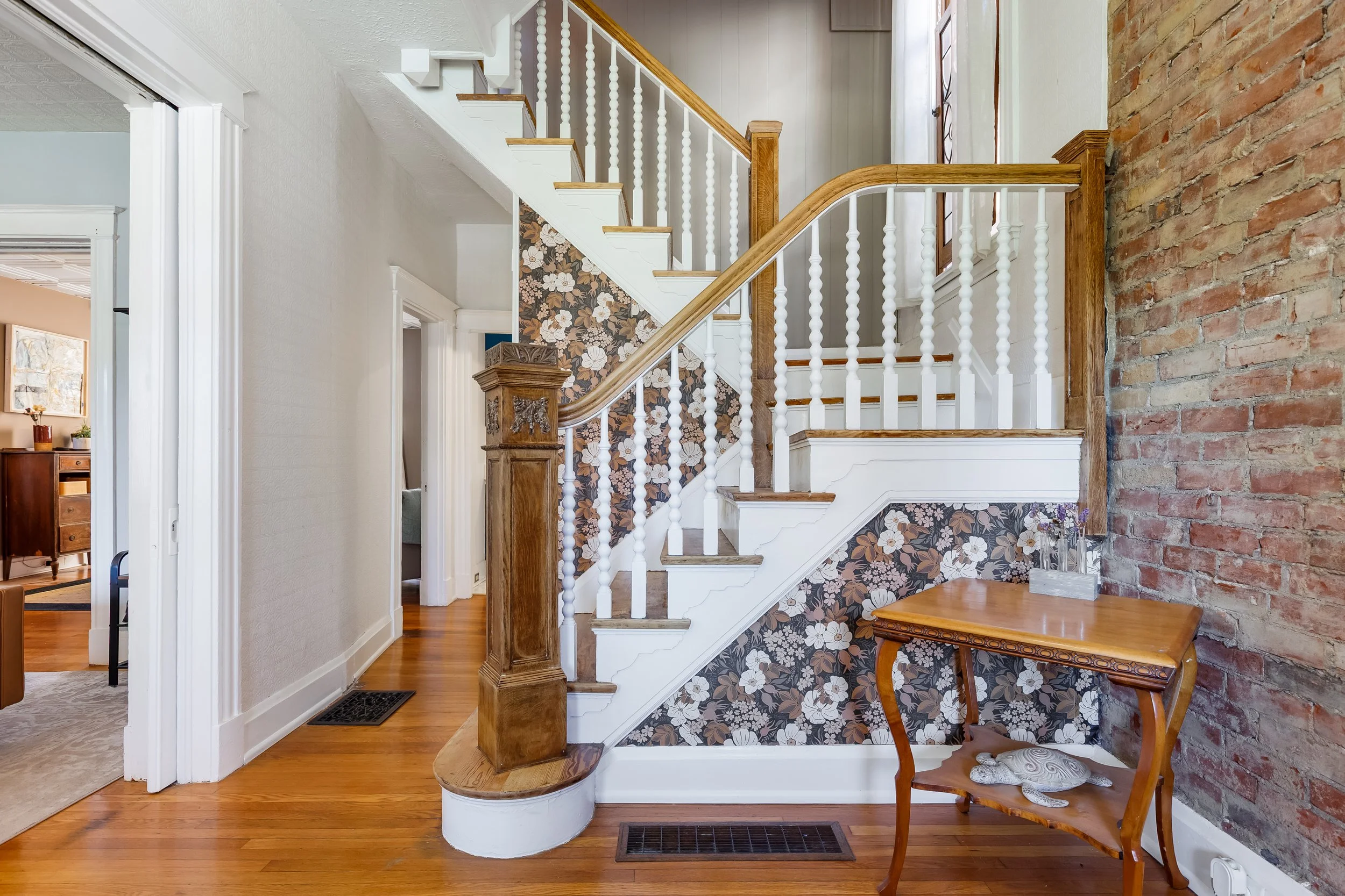 Interior view of a house showing a staircase with a floral wallpaper underneath, a brick wall to the right, hardwood flooring, part of a wooden table with a decorative object, and a doorway leading to other rooms.