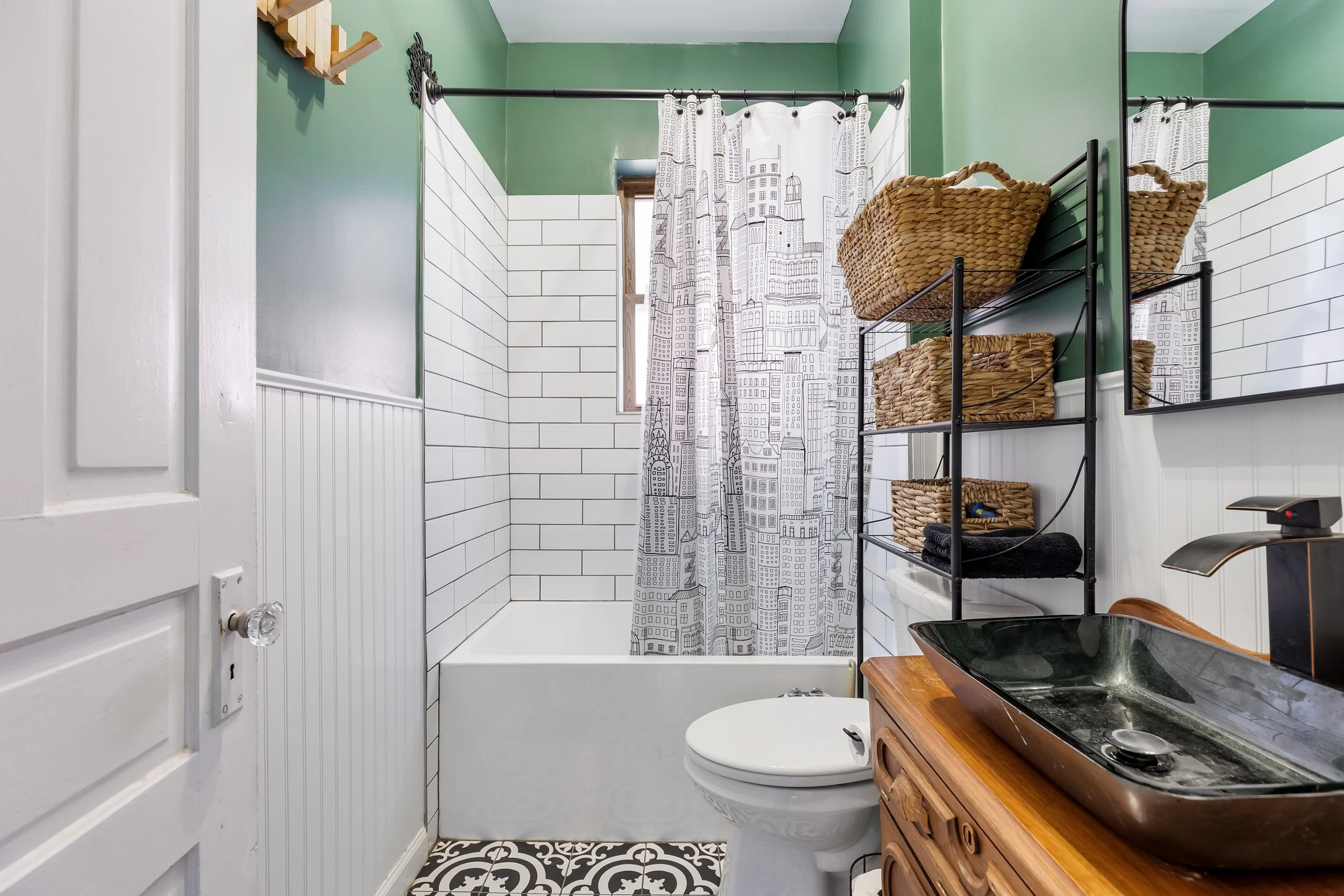 A small bathroom with green walls, white subway tile halfway up, and black and white patterned floor tiles. There is a shower with a cityscape themed curtain, a wooden vanity with a black vessel sink, a mirror, and a shelving unit with baskets.