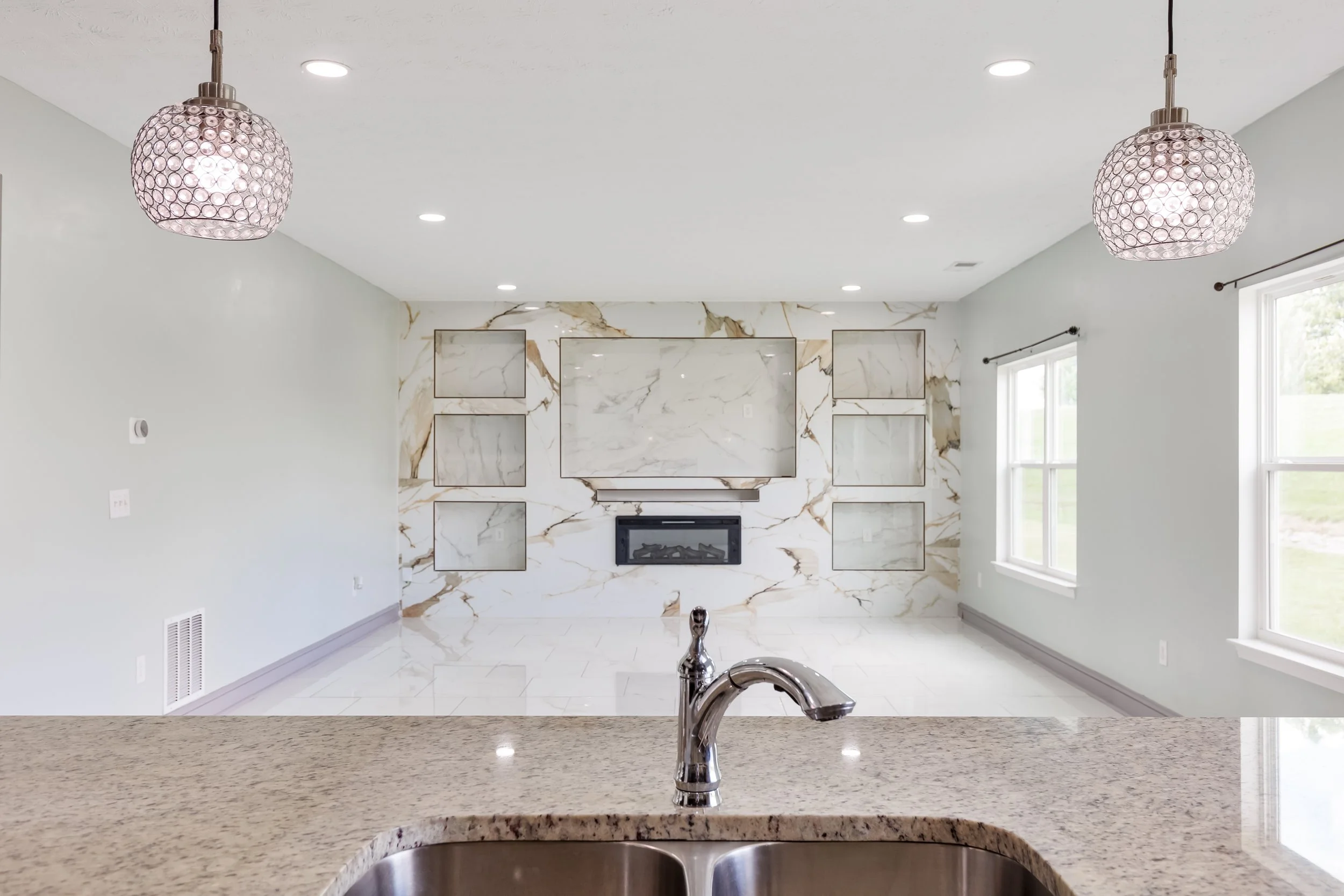 View of a modern living room with white marble fireplace, large windows, and a light-colored ceiling with recessed lighting.