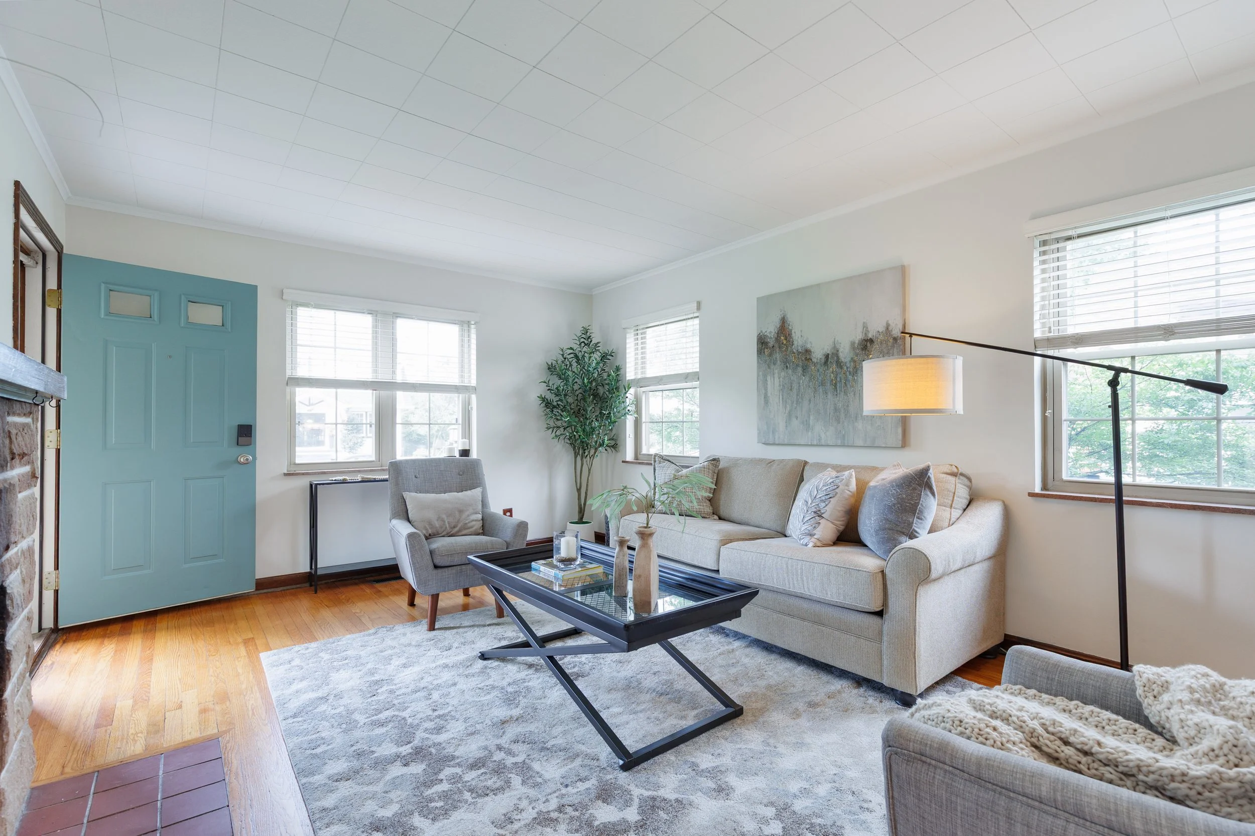 Living room with beige sofa, gray armchair, black coffee table, large windows, green plant, abstract painting, floor lamp, and hardwood floor with gray rug.