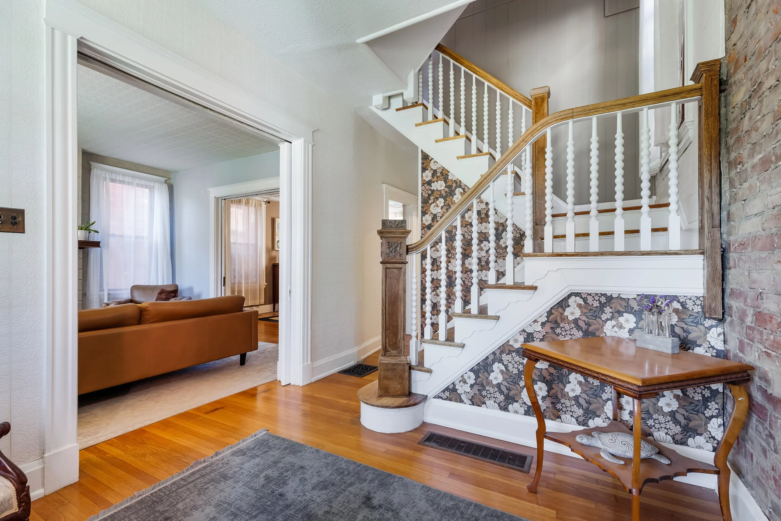 A view of a living room and staircase with floral wallpaper, white railings, and wooden steps, with portions of brick wall on the right and windows with sheer curtains in the background.