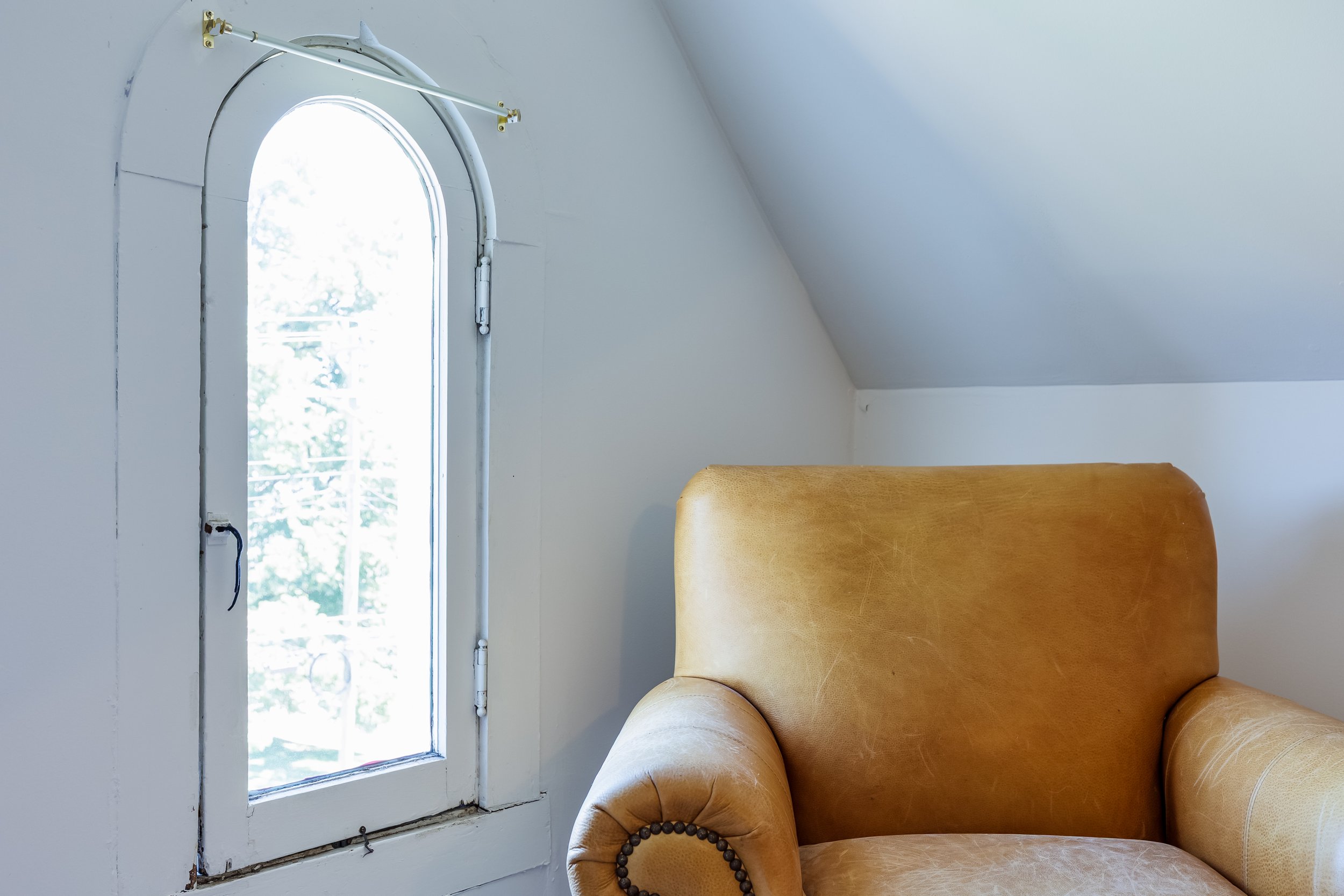 A partially opened window with a white frame and a view of trees outside, next to a mustard-colored leather armchair in a room with white walls and a sloped ceiling.