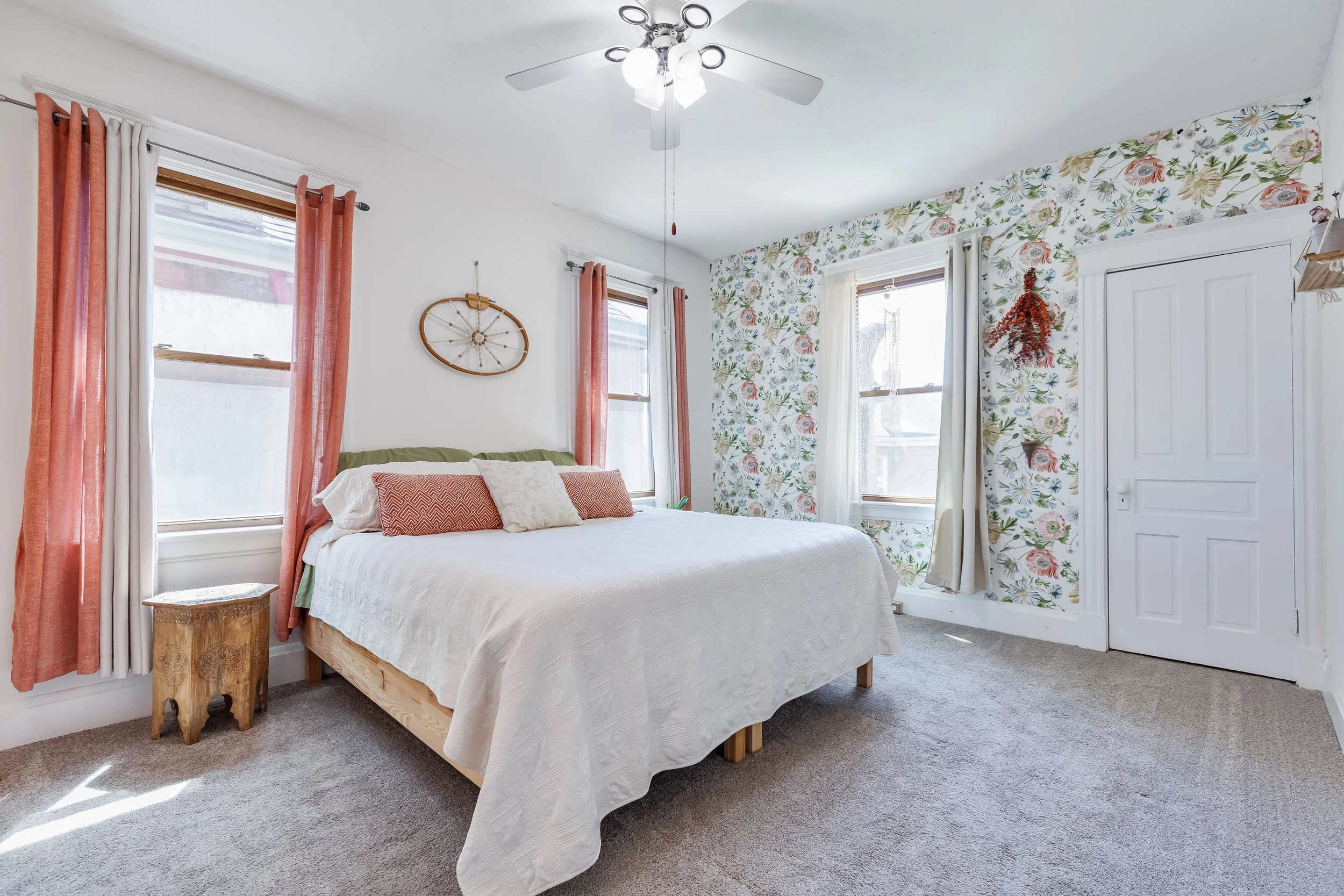 A bedroom with a white ceiling fan, two windows with white and coral curtains, a bed with a white quilt and decorative pillows, a wooden nightstand, and a floral wallpaper accent wall.