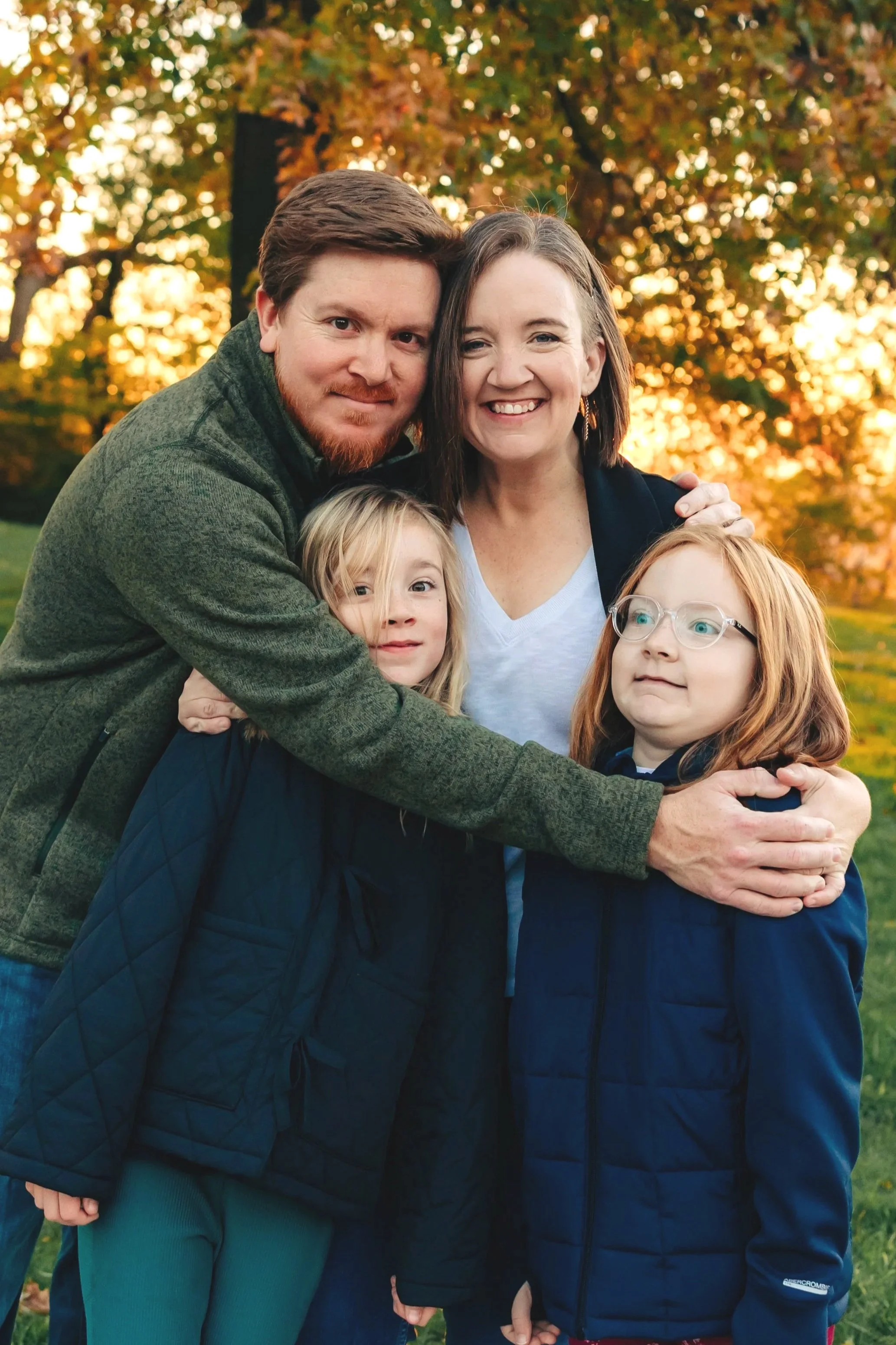 A family of five standing outdoors in front of autumn trees. The adults are smiling and hugging. The children have neutral expressions, one with glasses and red hair, and the other with blonde hair.