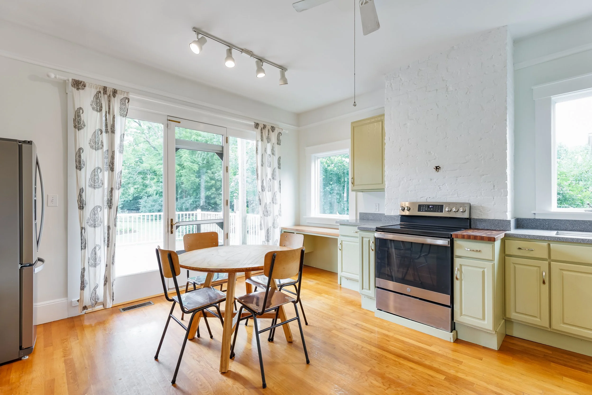 A bright kitchen with wooden floors, white walls, and large windows with patterned curtains, featuring a round wooden dining table with four chairs, a stainless steel stove, and pastel yellow cabinets.