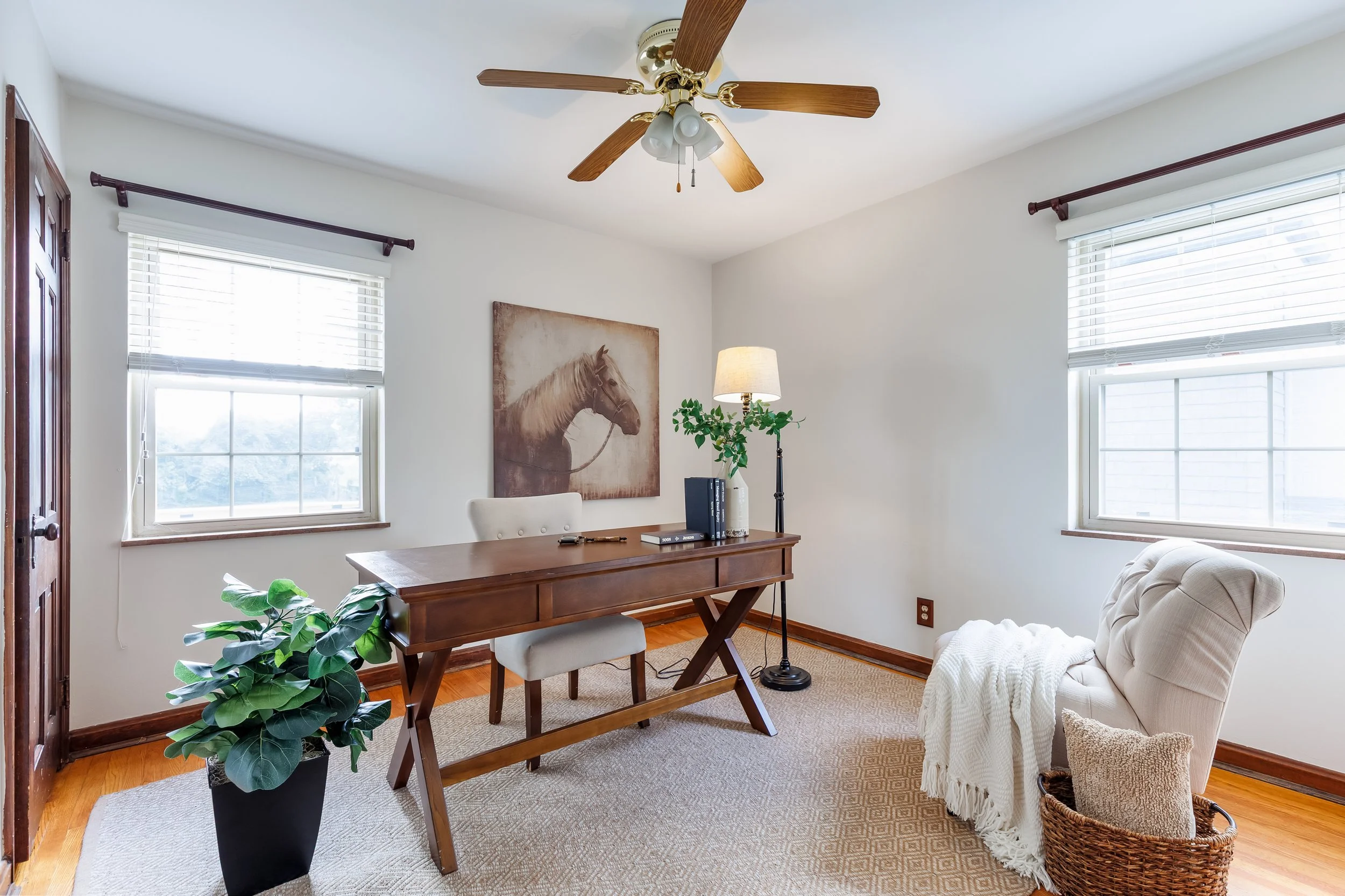 Living room with white walls, two large windows with blinds, a wooden desk with a white chair, a floor lamp, framed horse artwork, a beige armchair with a white blanket, a wicker basket, and a potted plant.
