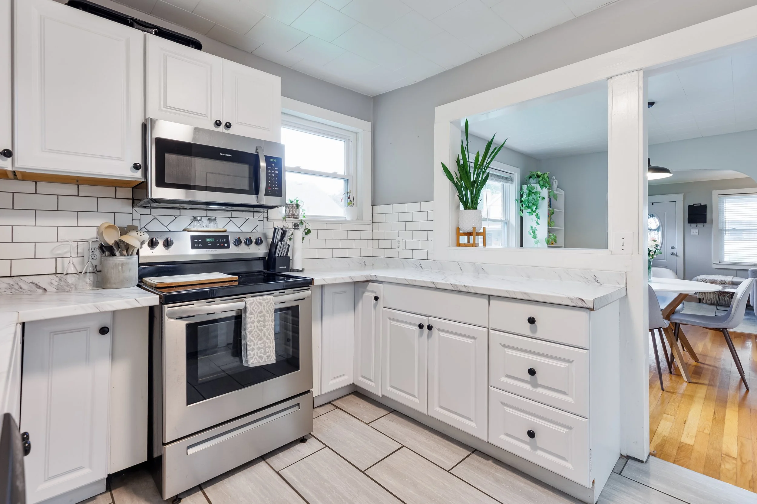 A bright kitchen with white cabinets, a stainless steel microwave and oven, white tiled backsplash, marble countertops, and plants near the windows.
