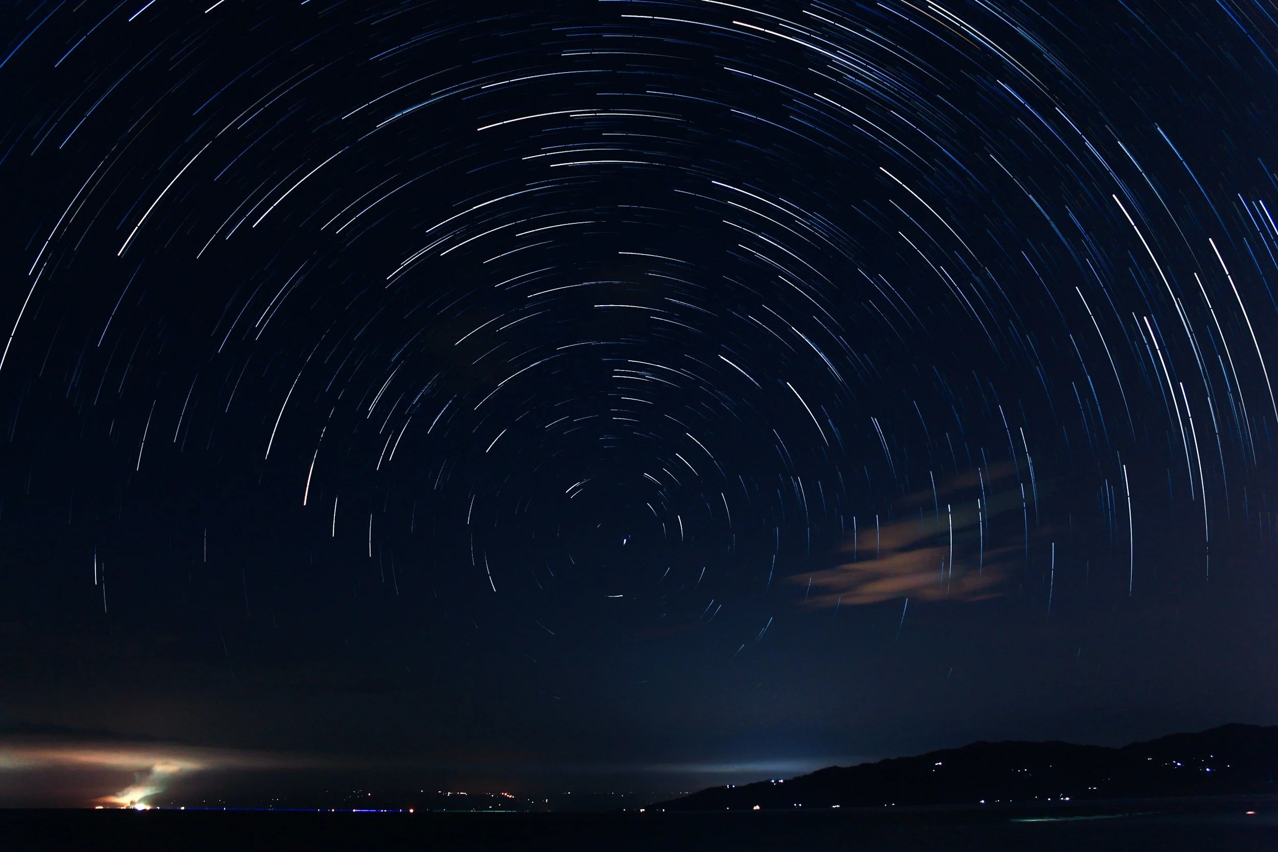 Night sky with star trails forming circular patterns above a night landscape with water and distant mountains.
