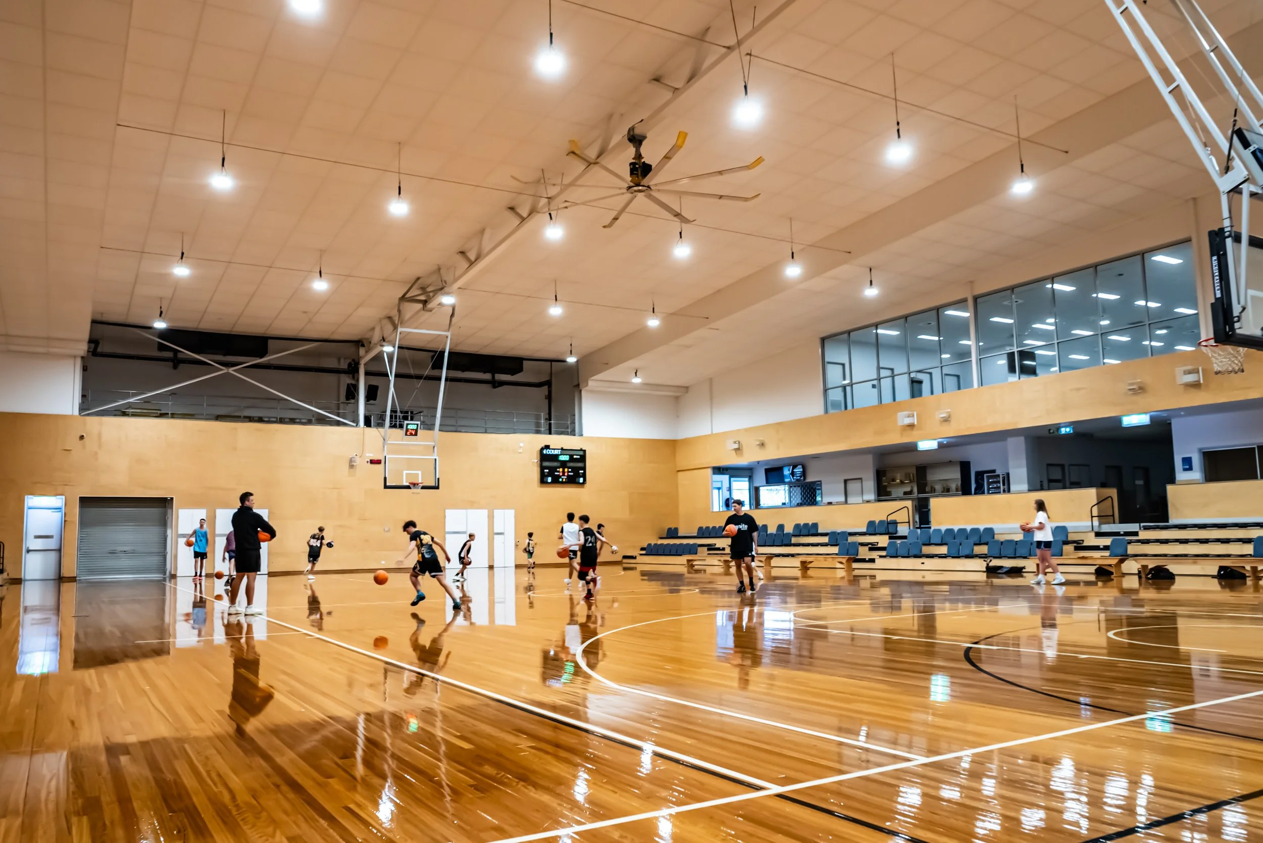 Indoor basketball court with children and adults playing. The court has a polished wooden floor, basketball hoops, and an electronic scoreboard. There are seating areas along the side and large windows on the upper level.
