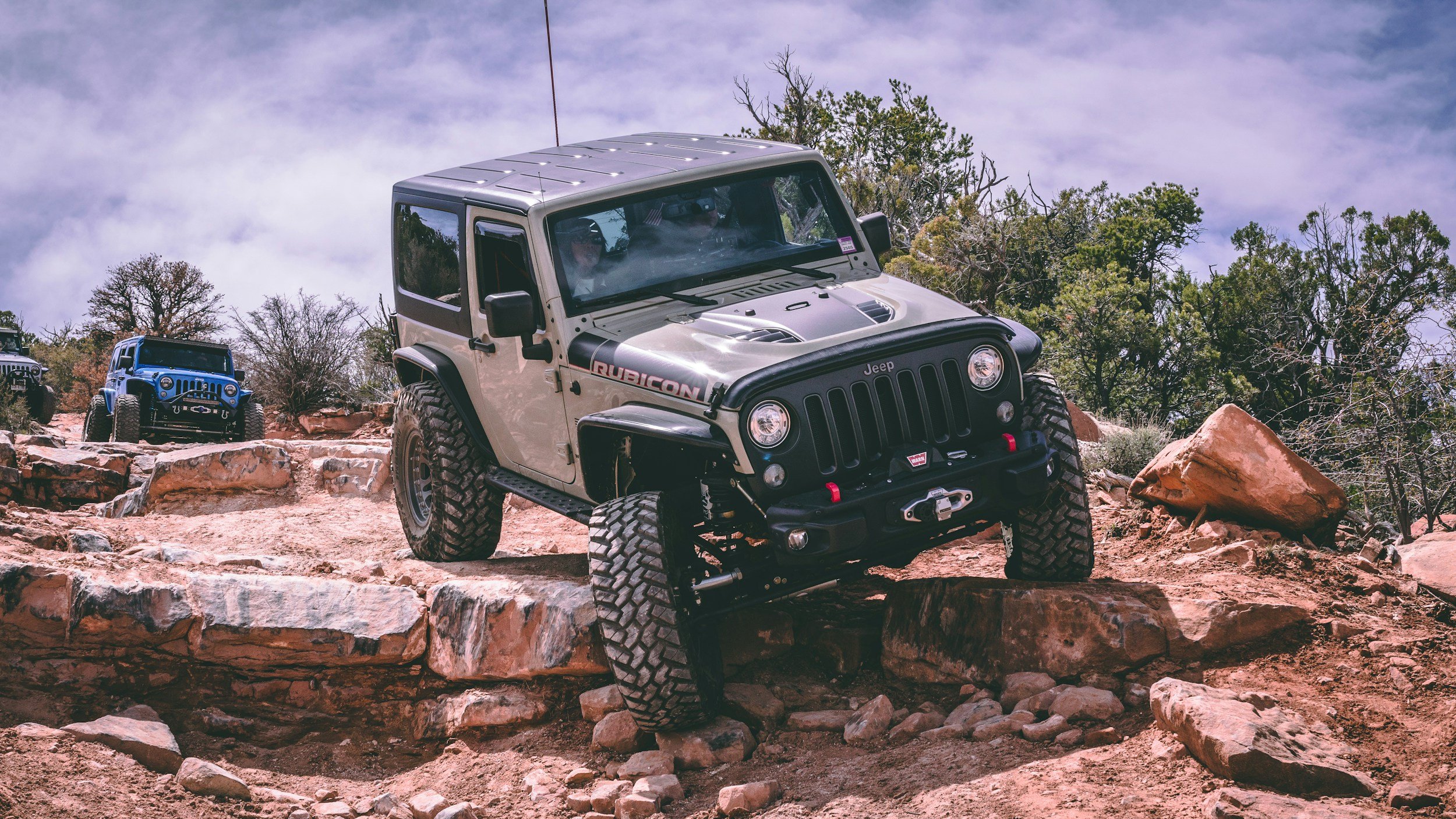 Two Jeep vehicles driving on rocky terrain in a desert landscape with trees and mountains in the background.