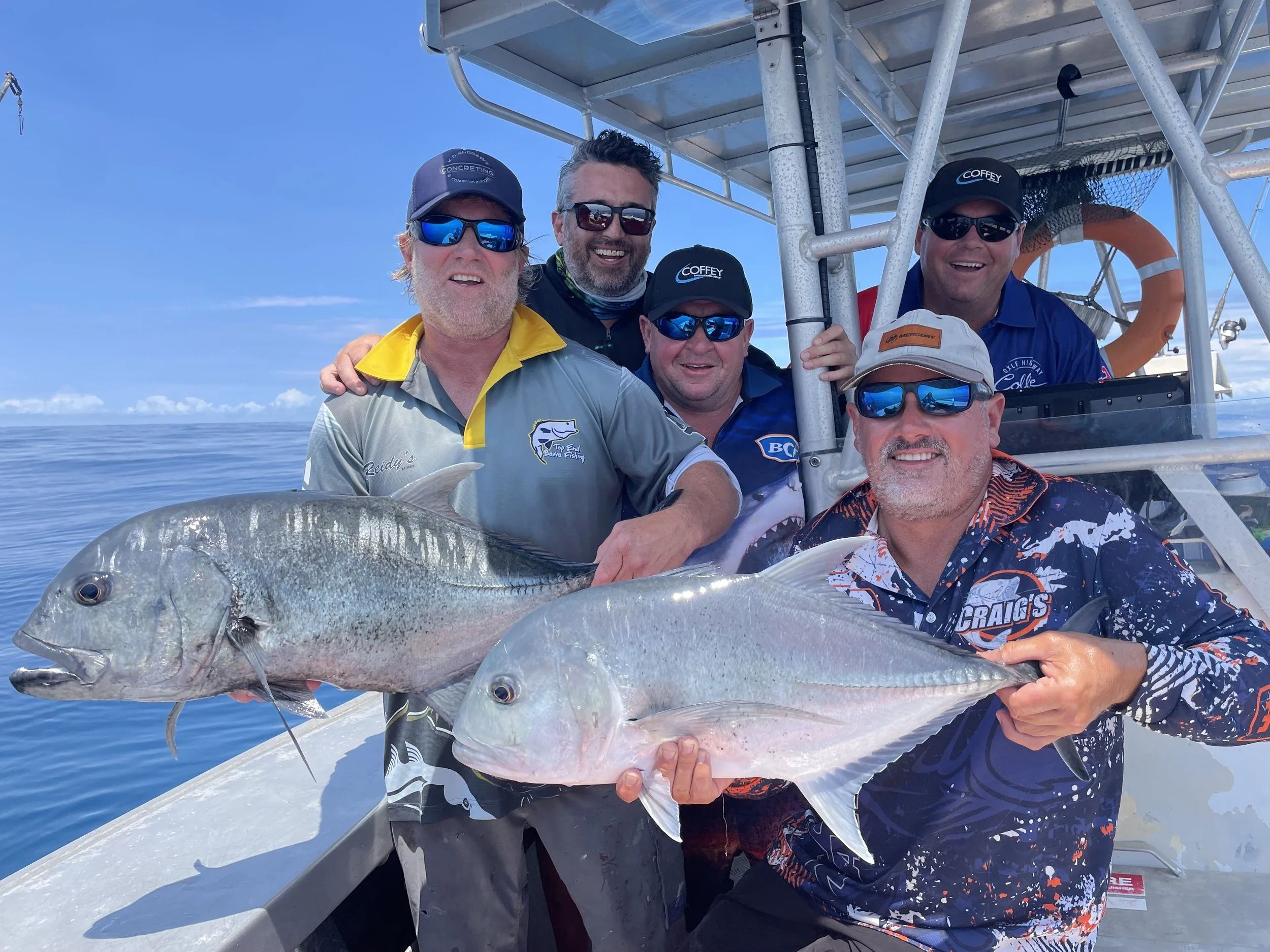 A group of five men on a boat holding two large fish, with a clear blue sky and calm ocean in the background.