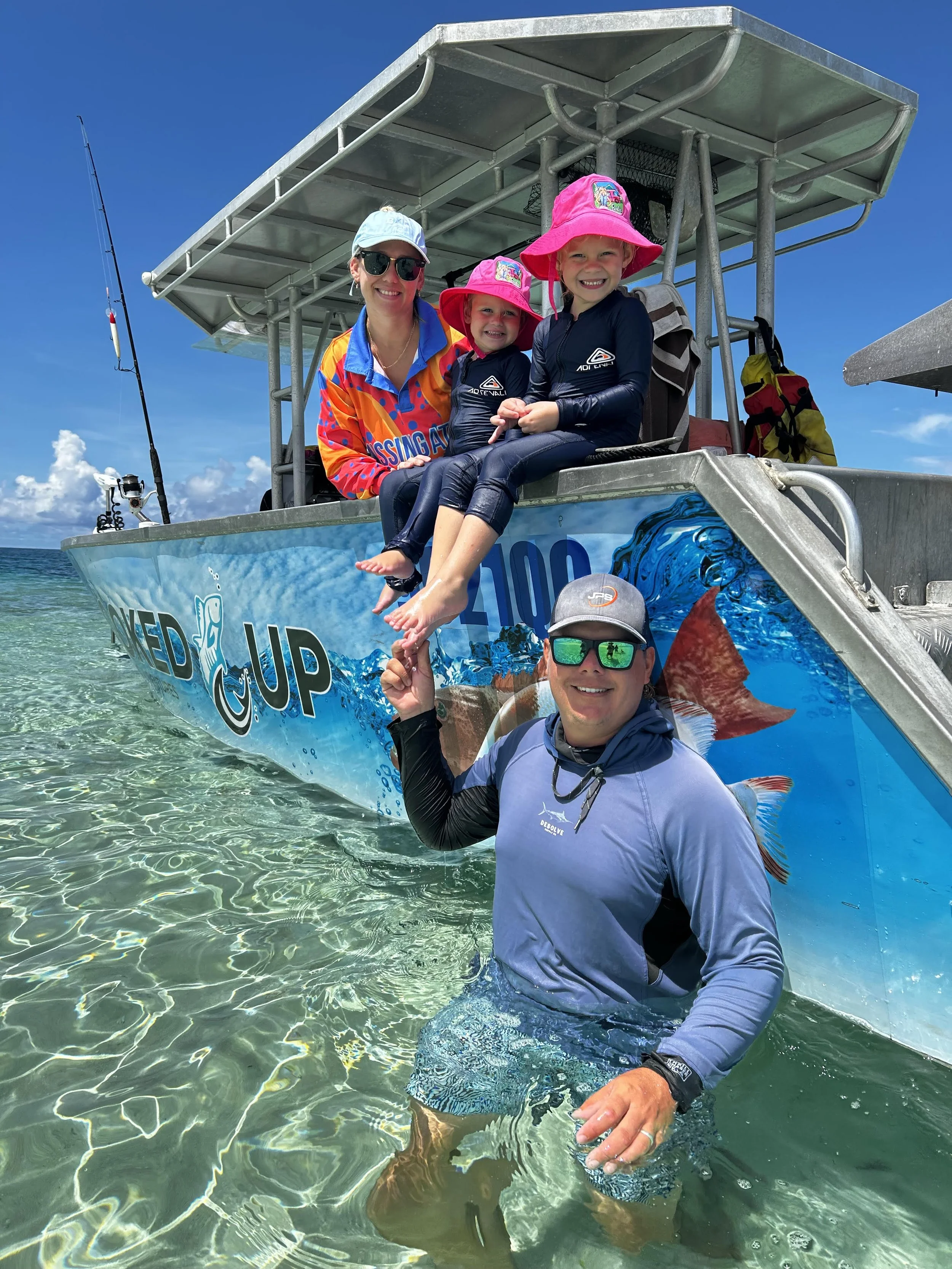 A group of four people, three children and one adult, posing on a boat in clear, shallow water. The children are sitting on the edge of the boat wearing colorful hats and wetsuits, smiling. The adult is standing in the water, holding the children's feet, wearing sunglasses and a long-sleeve shirt.
