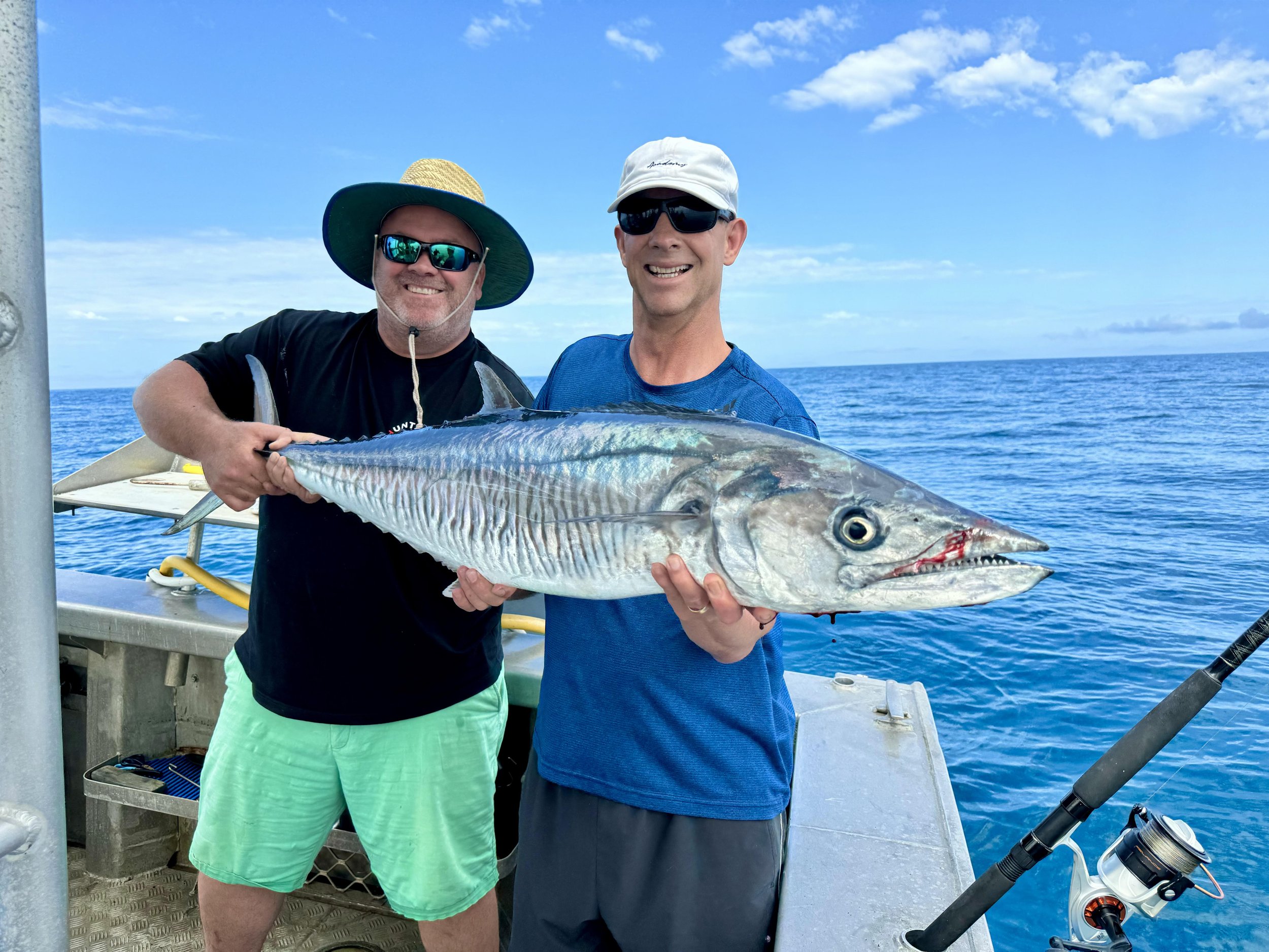Two men on a boat holding a large fish, likely a mackerel or similar species, with a blue ocean and sky in the background.