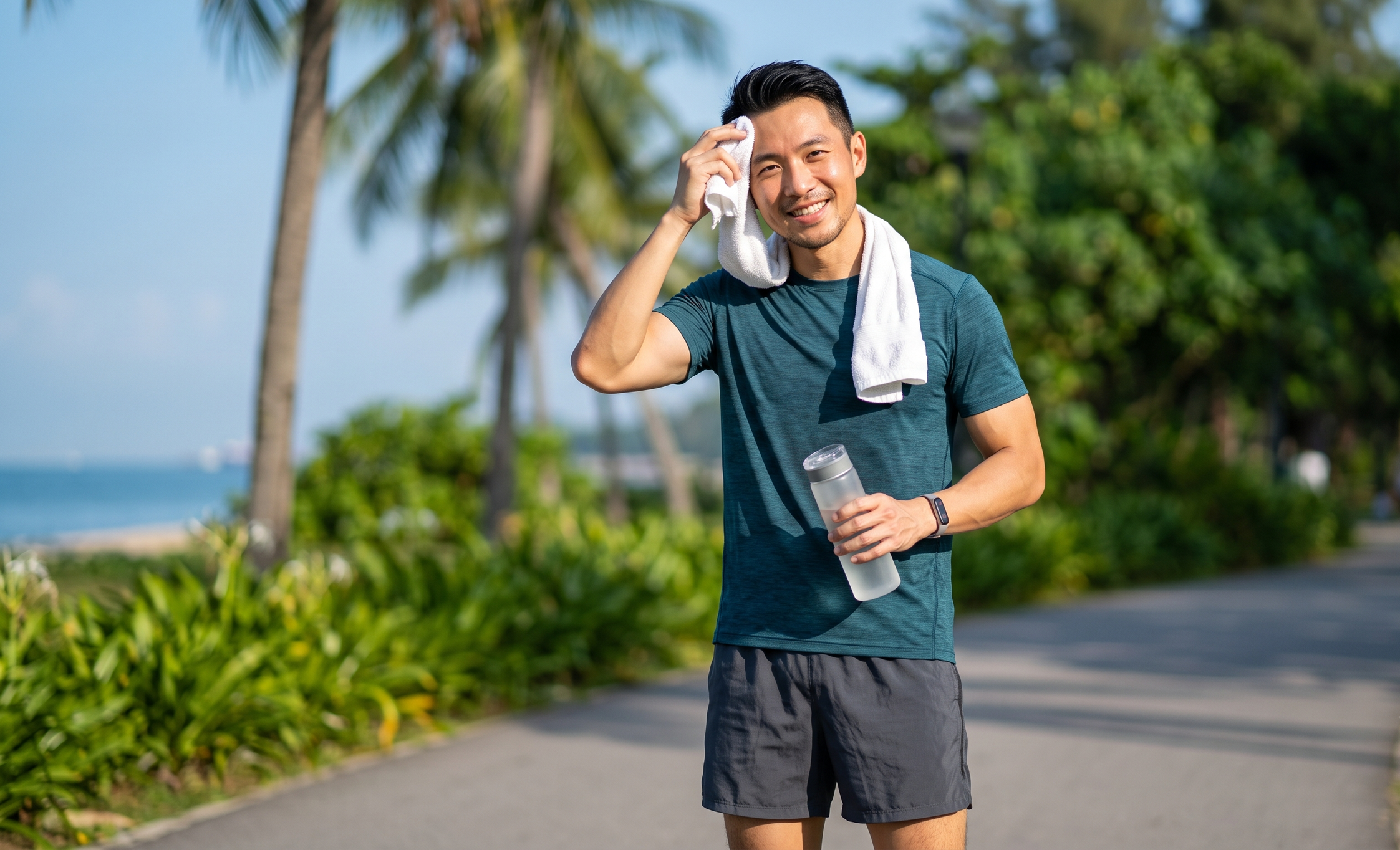 A young man with a towel around his neck, holding a water bottle, smiling after jogging outdoors on a sunny day near trees and greenery.