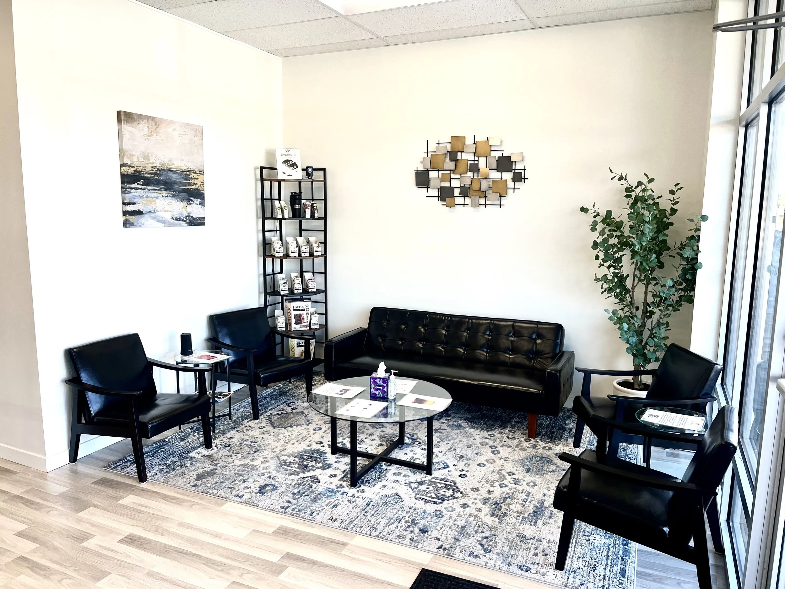 Waiting room with four black armchairs, a black leather sofa, a round glass coffee table, a tall black shelf with magazines and books, a large side window, a green potted plant, wall art, and a patterned area rug.