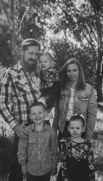 Family of five outdoors, two adults and three children, under trees.