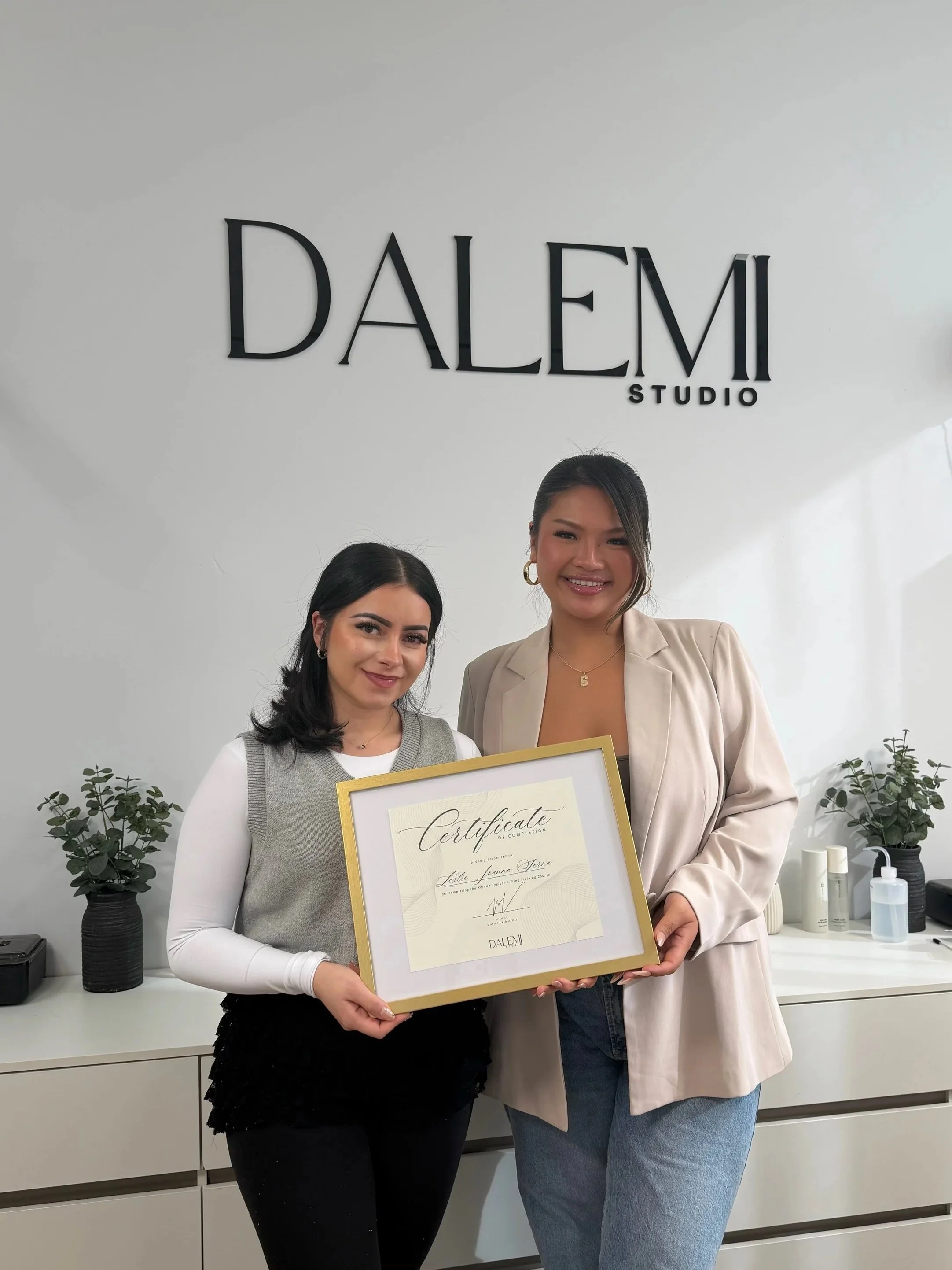 Two women smiling and holding a large certificate in front of a white wall with 'DALEM STUDIO' sign. There are plants and skincare bottles on the counter behind them.
