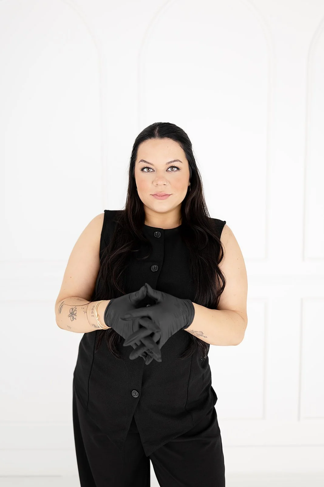 A woman with long dark hair, wearing a sleeveless black dress and black gloves, standing against a white background.