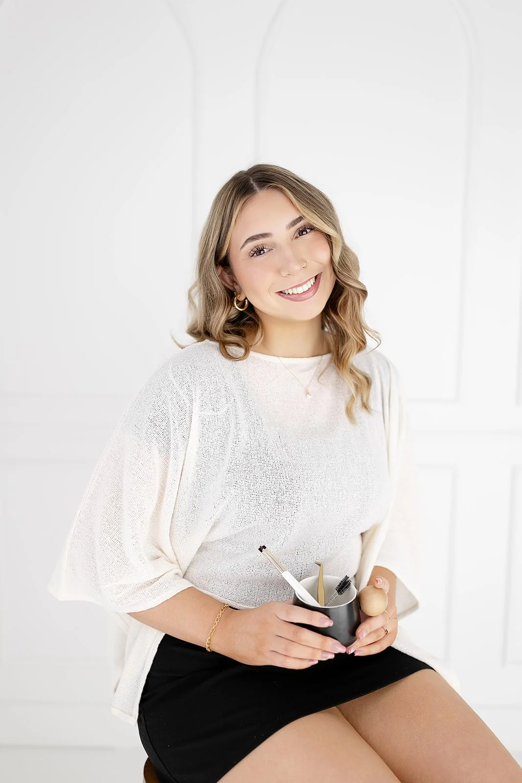 Smiling woman with wavy light brown hair, wearing a white knit top, black skirt, and gold jewelry, sitting against a white background and holding a black mug filled with various beauty tools.