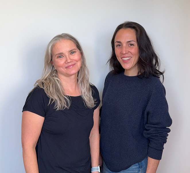 Two women standing close together, smiling, against a plain white wall.