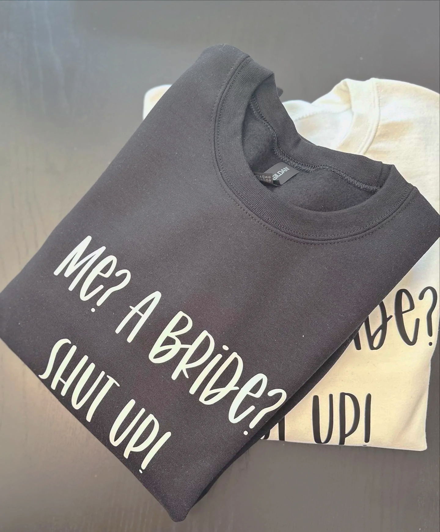 Folded black T-shirt with the humorous text 'MRS? A BRIDE? SHUT UP!' printed in white, placed on top of a white T-shirt with the same text partially visible.