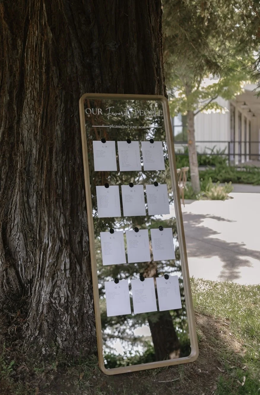 A mirror tree-shaped seating chart stand outdoors, displaying sheets of paper with seating arrangements for a wedding or event, with trees and a building in the background and custom vinyl lettering.