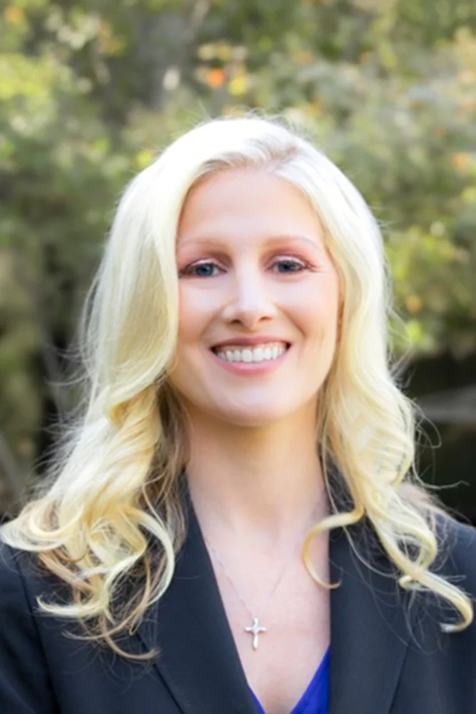 Tara Tobin, CPA, MSTwearing a black blazer and a small silver cross pendant, outdoors with blurred trees in the background.