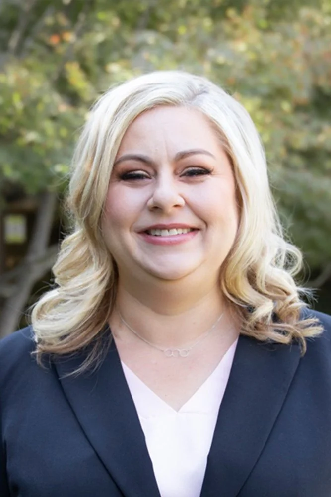 Kristin Monsibais, CPA, MST wearing a navy blazer and white top, standing outdoors with trees in the background.
