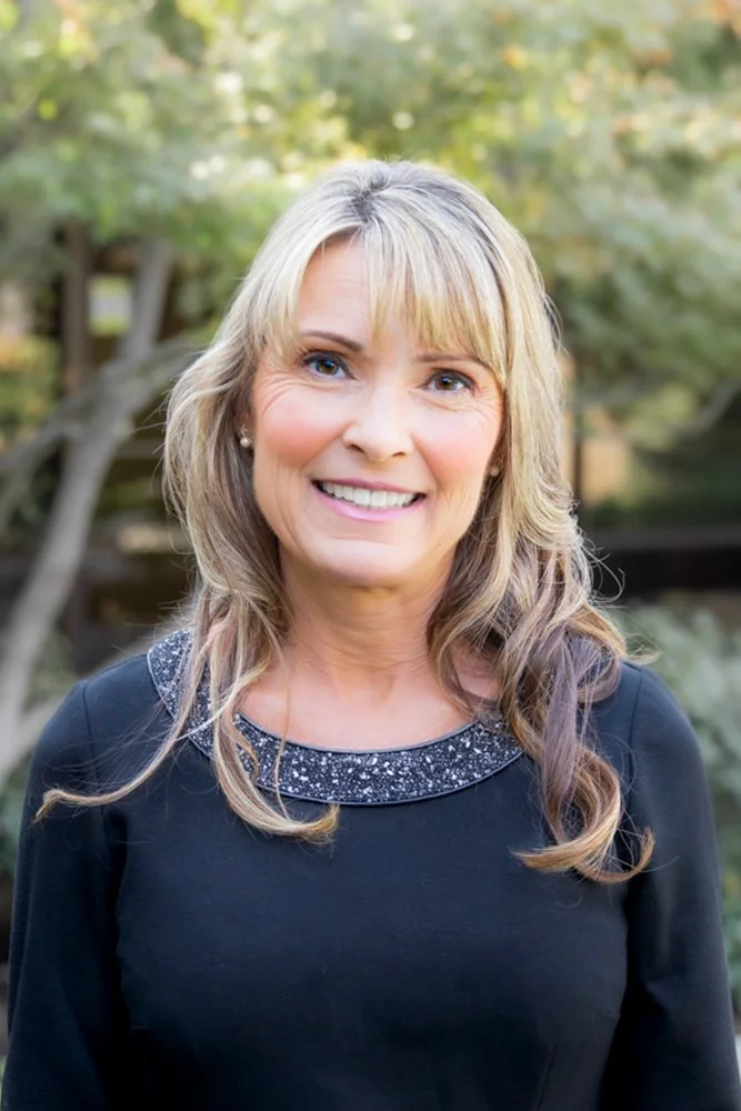 Valerie Adams, CPA, MST, wearing a black top with a sparkly collar, standing outdoors with blurred trees in the background.