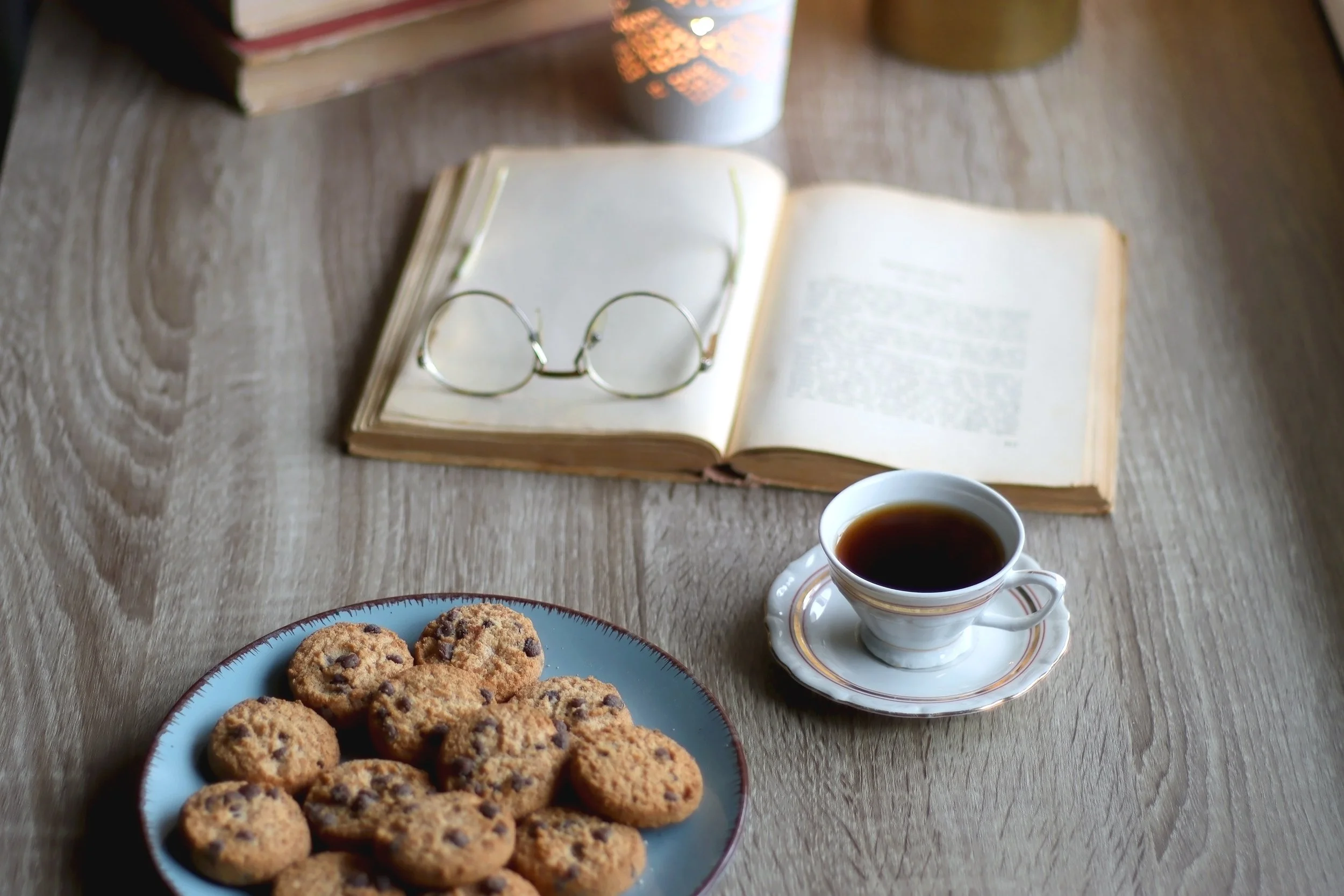 A wooden table with an open book, a pair of eyeglasses on the book, a cup of black coffee in a saucer, and a plate of chocolate chip cookies. In the background, there are some stacked books and a candle.