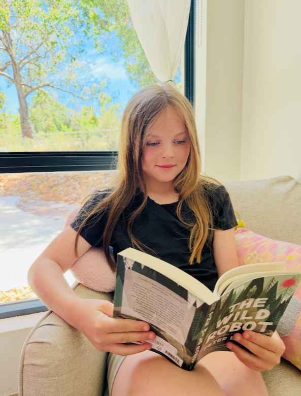 A young girl with long brown hair is sitting on a beige couch, reading a book titled 'The Wild Robot'. She is wearing a black T-shirt and shorts, and is near a large window with a view of tree branches and blue sky outside.