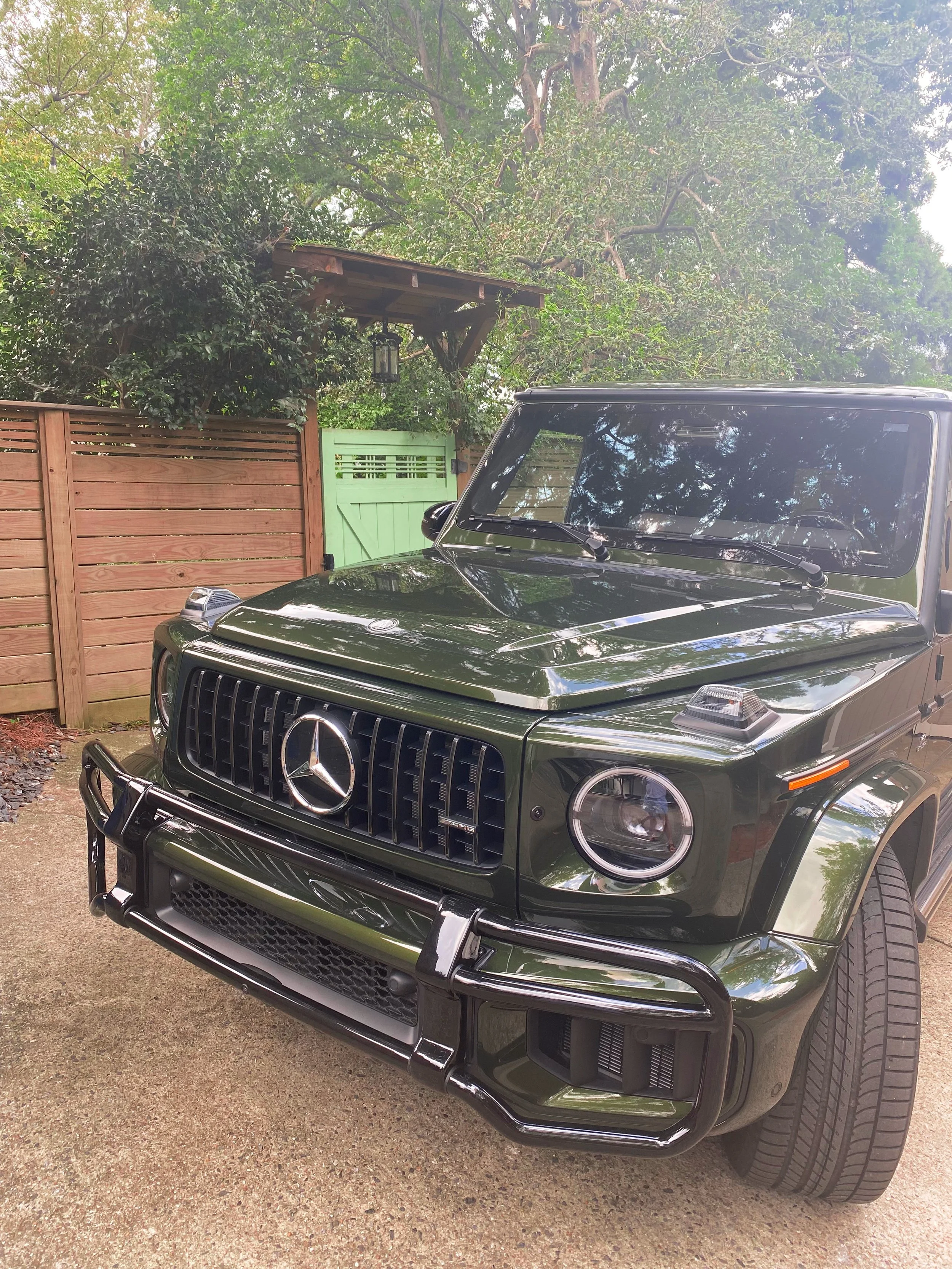 An olive green 2025 Mercedes-Benz G63 SUV parked outdoors on a driveway, with greenery and a wooden fence in the background.
